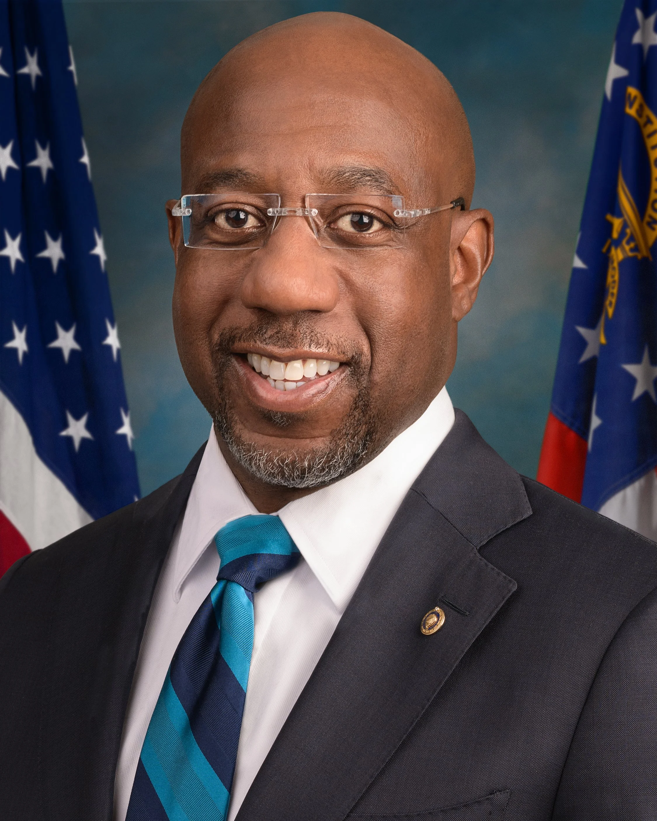 Close-up portrait of an African American man in a suit, wearing glasses, smiling, with an American flag and another flag in the background.
