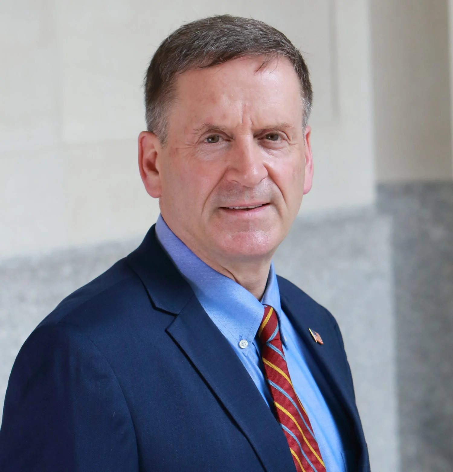 A middle-aged man with short brown hair and light skin, wearing a dark suit, blue shirt, and a patterned tie, standing indoors against a light-colored wall.