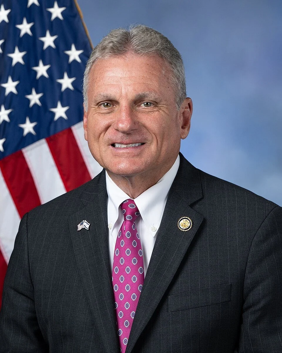 Portrait of a man in a dark suit with a purple patterned tie, positioned in front of an American flag.