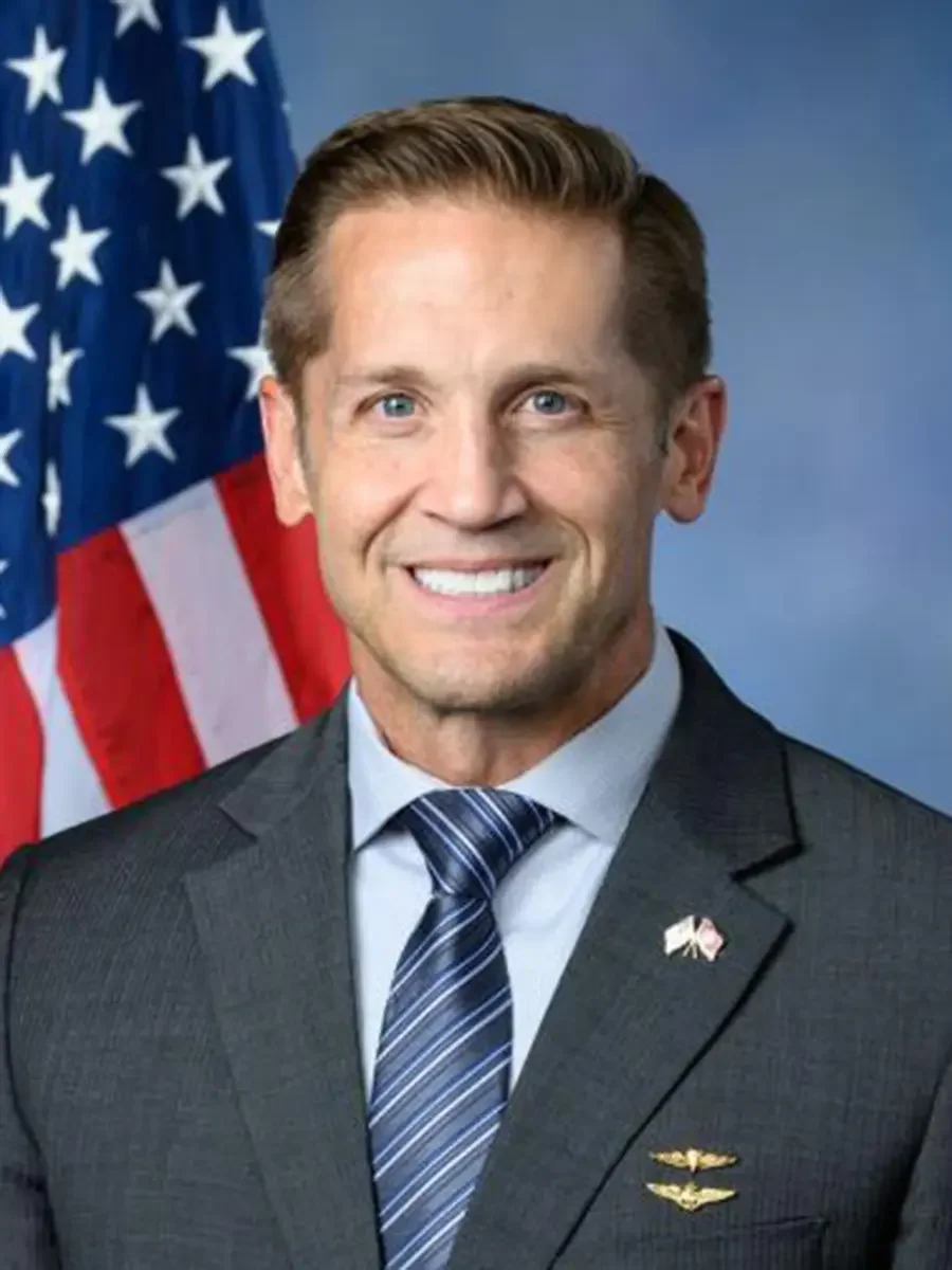 A smiling man in a dark suit, light blue shirt, and striped tie, standing in front of an American flag with a blue background.