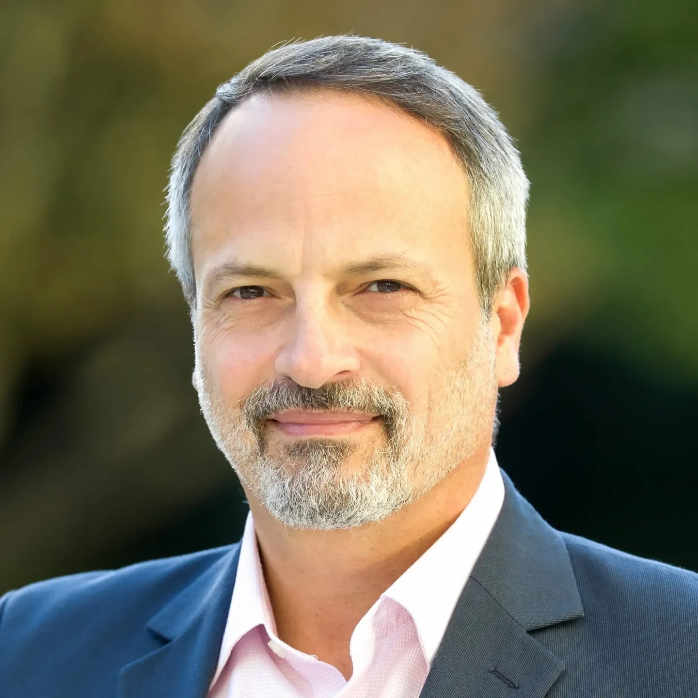 Close-up of a middle-aged man with gray hair, beard, wearing a suit jacket and pink shirt outdoors.