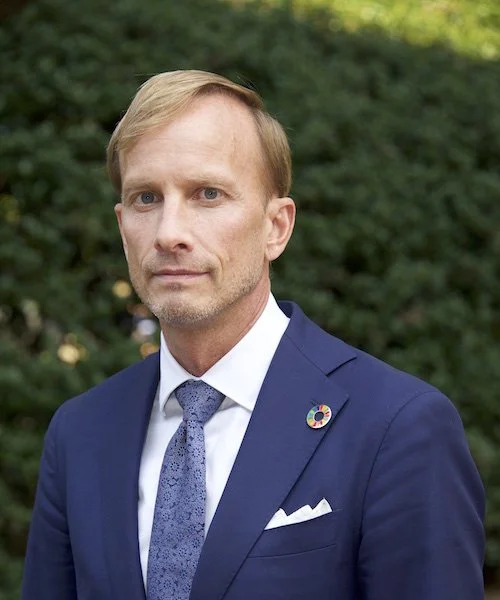 A man with blonde hair and a serious expression, wearing a dark blue suit, white shirt, patterned tie, and a lapel pin, standing outdoors with greenery in the background.
