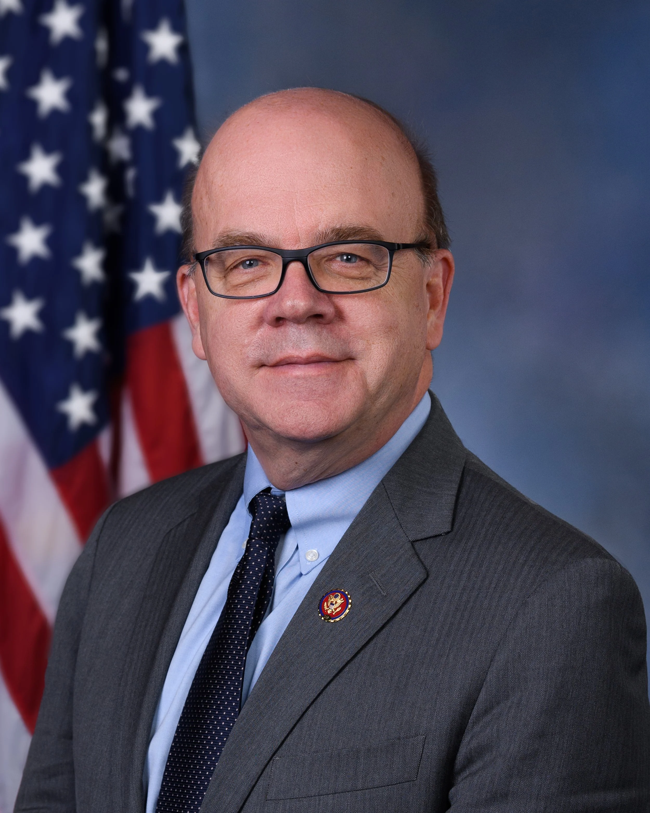 Close-up of a man with glasses, wearing a suit, standing in front of an American flag.