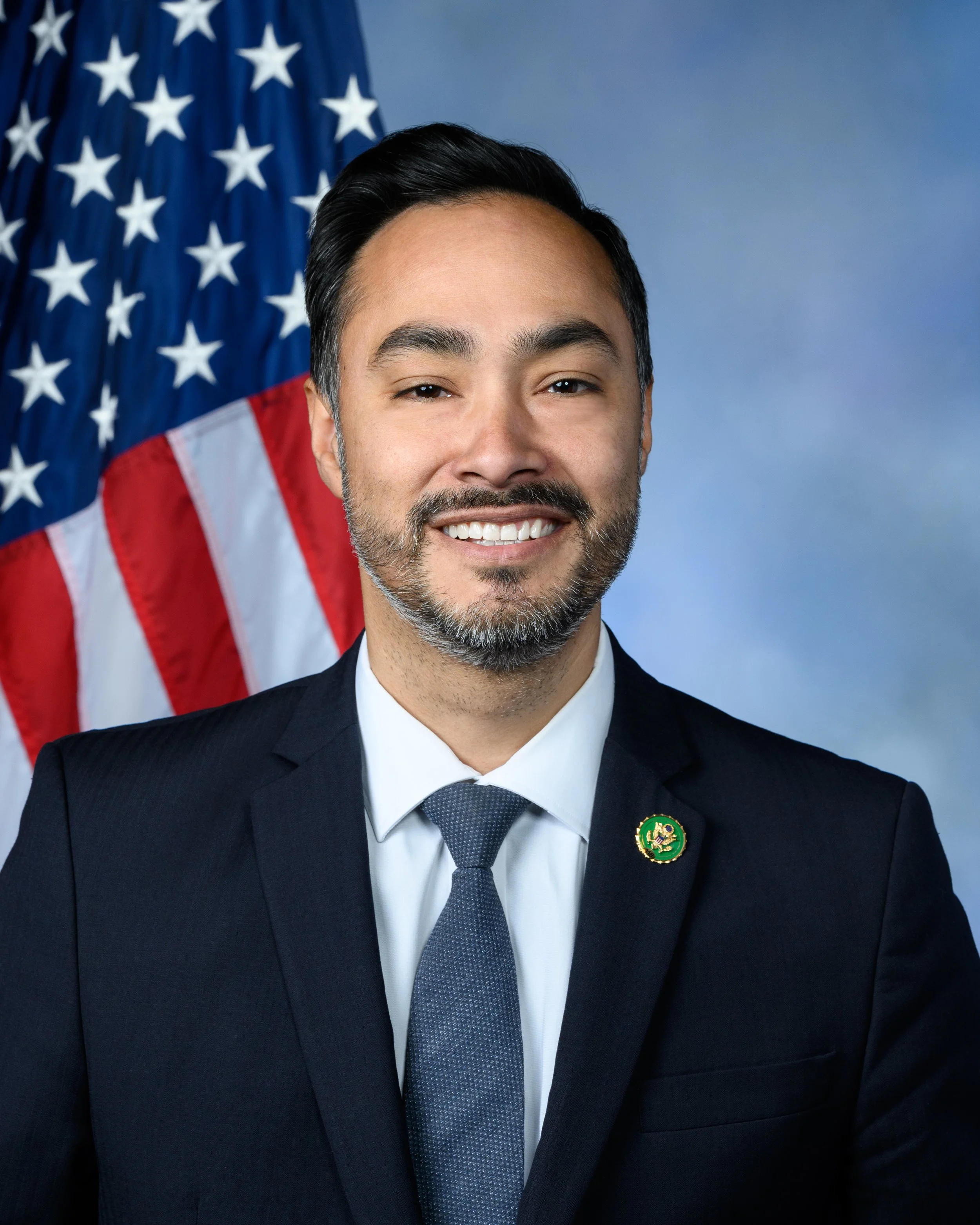 Close-up of a smiling man with dark hair and facial hair, wearing a dark suit, white shirt, and gray tie, standing in front of the American flag.