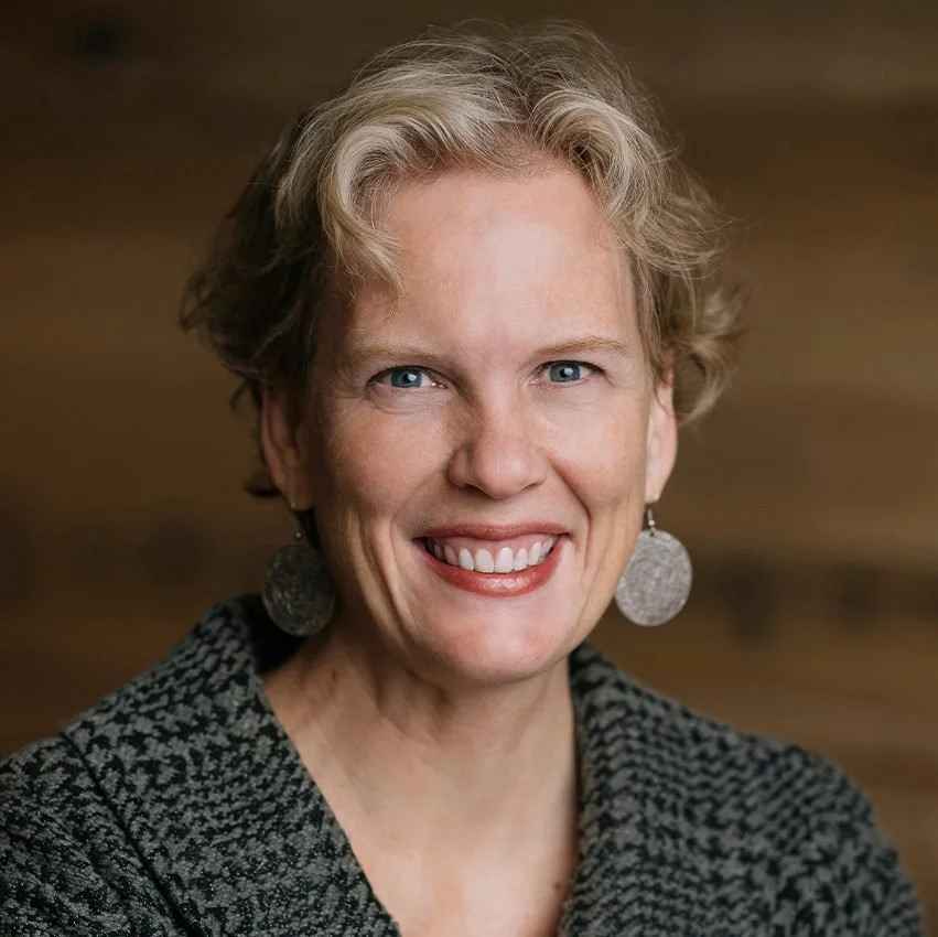 Close-up portrait of a smiling woman with short blonde hair, blue eyes, wearing large earrings and a patterned jacket, against a blurred wooden background.