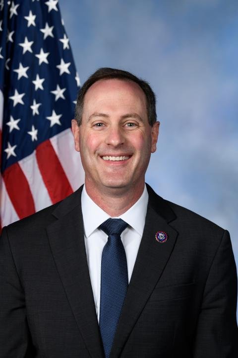 A smiling man in a black suit, white shirt, and blue tie standing in front of an American flag against a blue background.