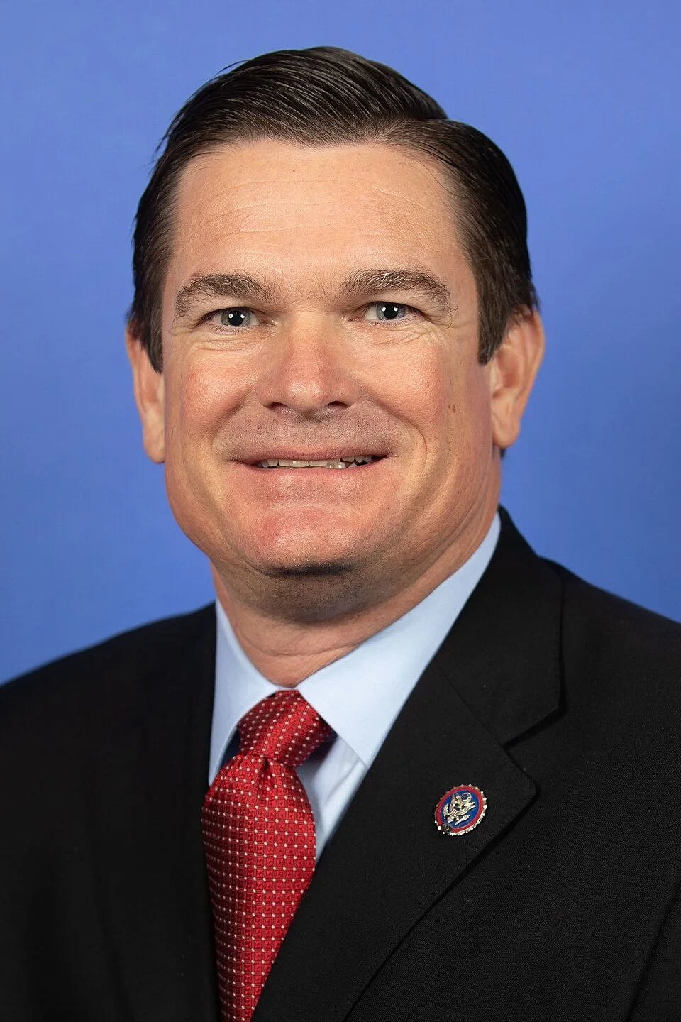 Headshot of a man in a suit with a United States lapel pin, smiling in front of a blue background.