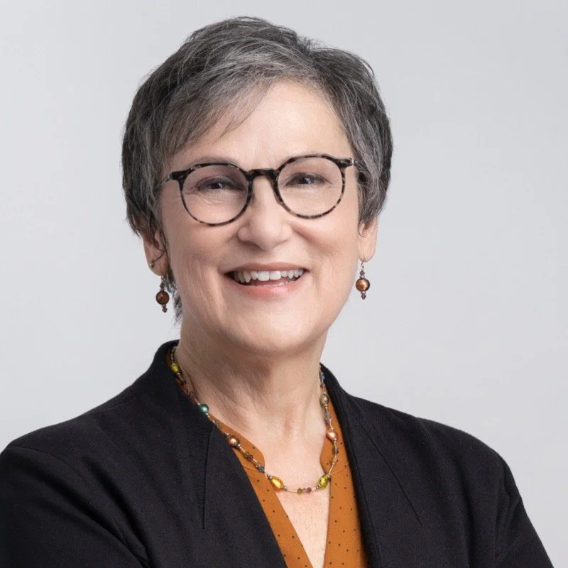 A middle-aged woman with short gray hair, glasses, and earrings, smiling at the camera. She is wearing a black blazer over an orange blouse with polka dots, and colorful jewelry, against a plain light gray background.