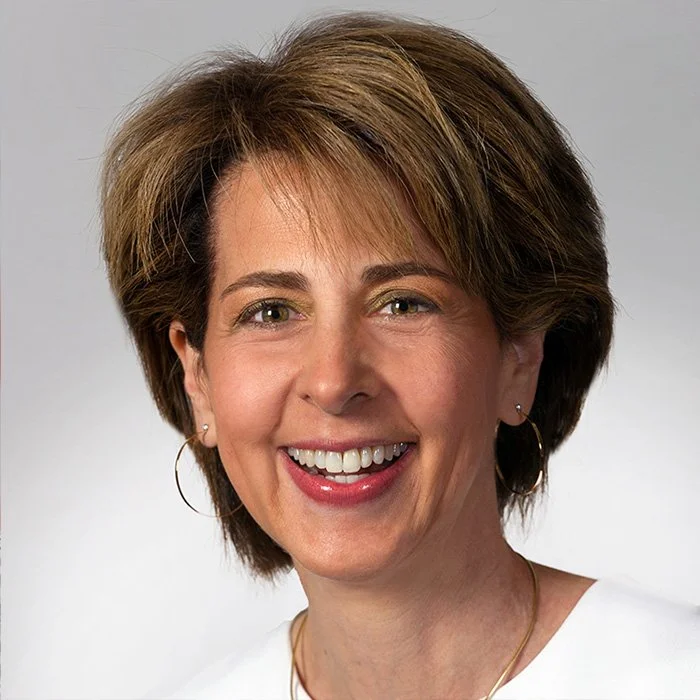 A close-up of a smiling woman with short brown hair, wearing hoop earrings and a white top, against a plain grey background.
