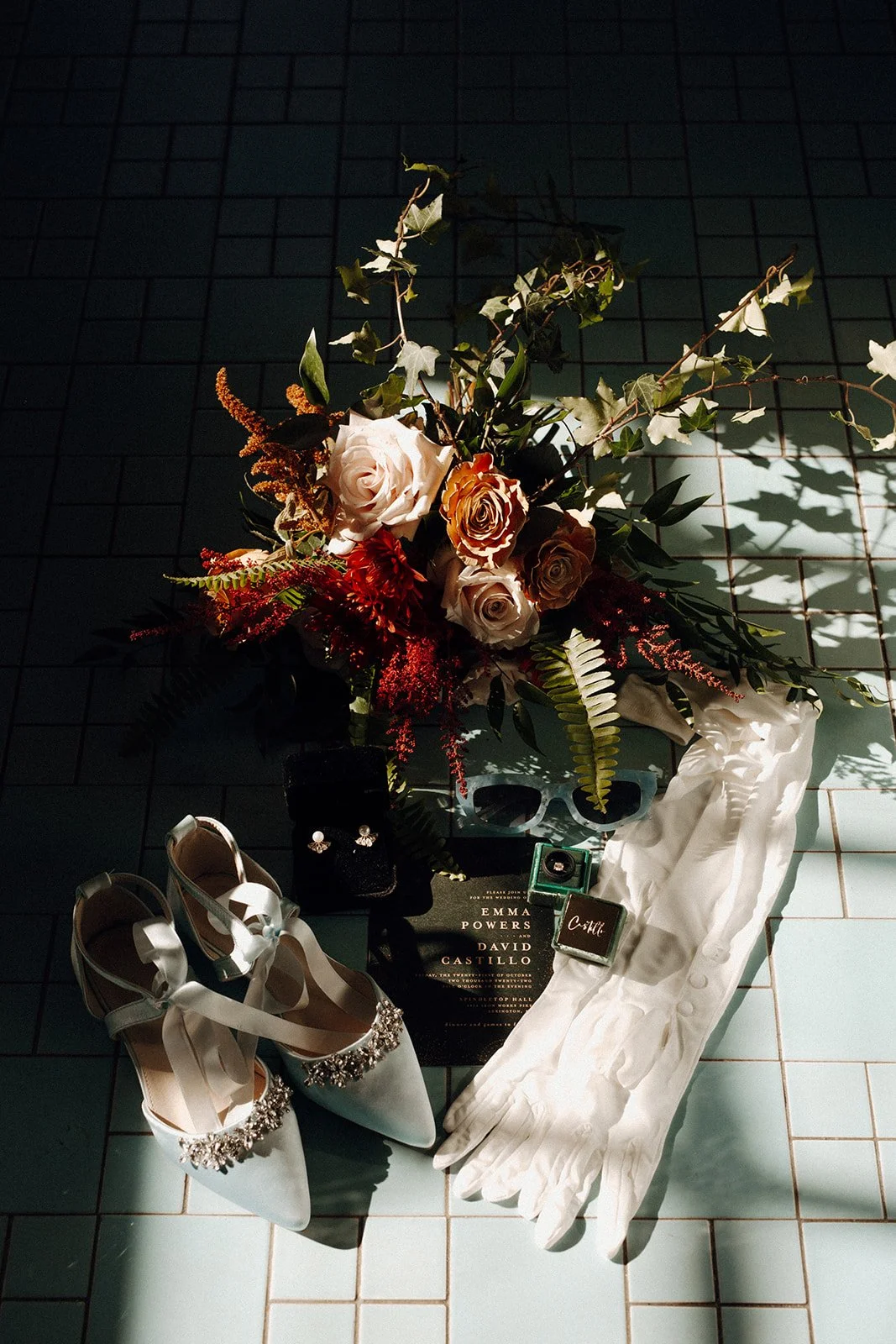 Still life of a wedding bouquet, long white gloves, white rhinestone heels, teal sunglasses, bride's ring and couple's printed invitation, against vintage baby blue tile.