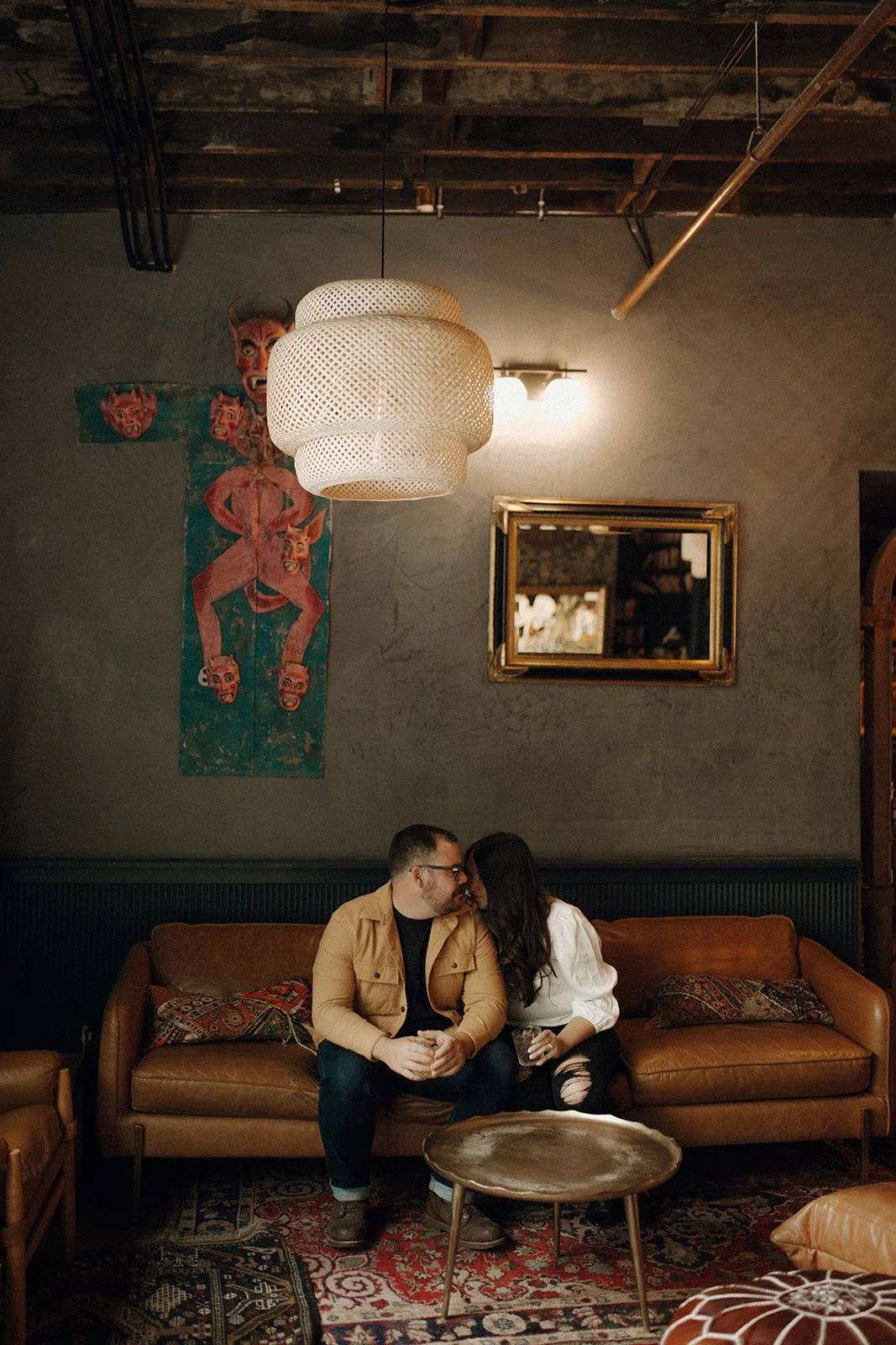 Sitting on camel leather couch in a living room with art and mirror on back wall, a couple touch faces in an intimate moment.