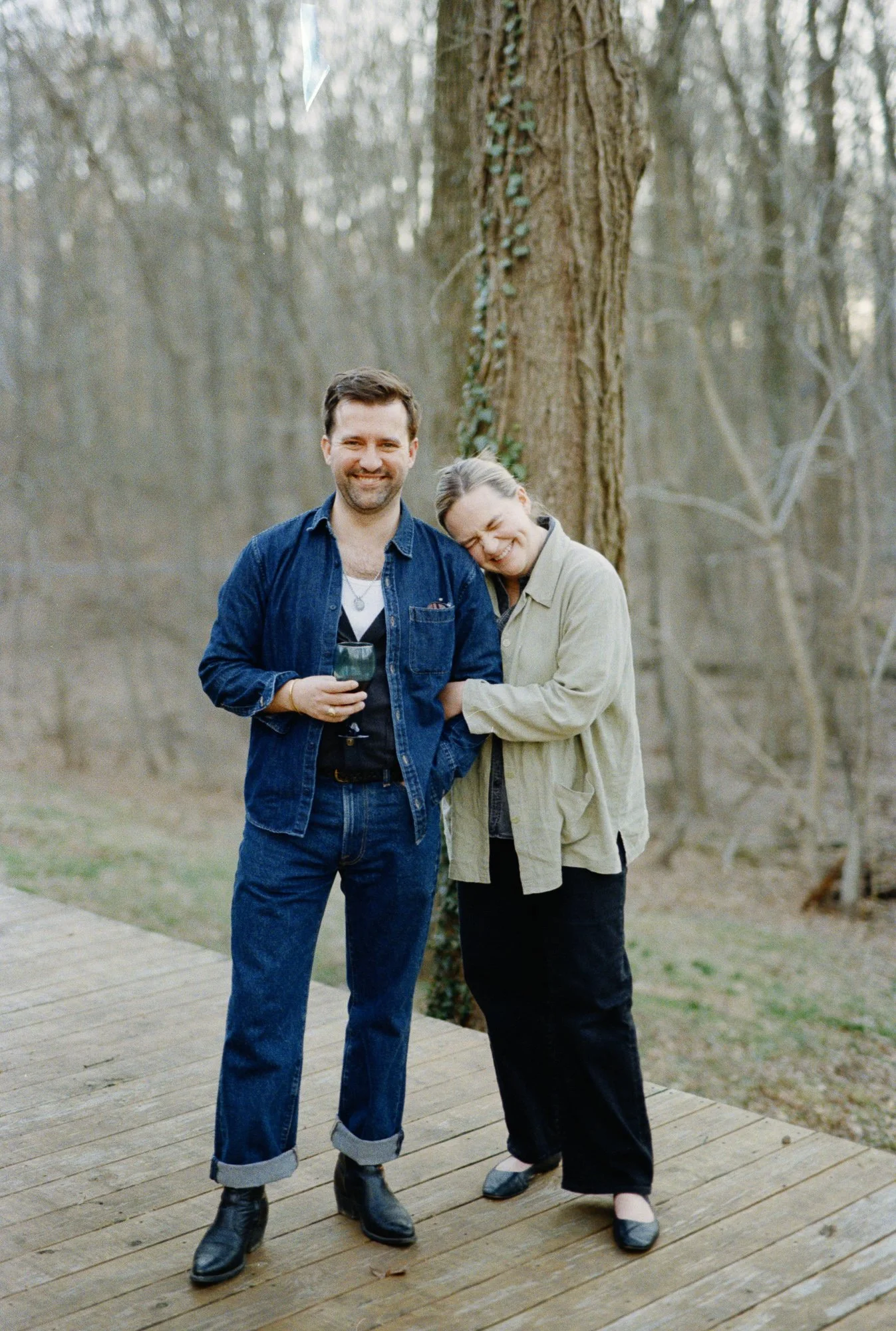 Couple standing on a wooden deck. Woman's arm is wrapped in Man's arm he is holding a glass and smiling and she has her head leaning on his shoulder while also smiling with her eyes closed