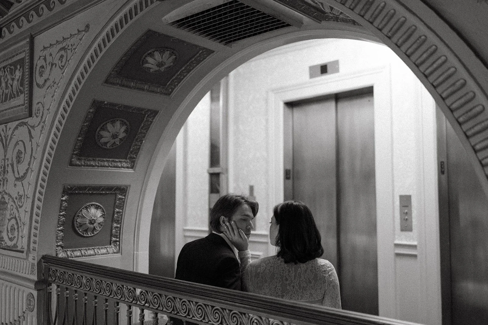 Black and white photo of a bride and groom in a private moment sitting on bench outside elevator inside decorative building with wood carvings on arched cutout and wall paneling