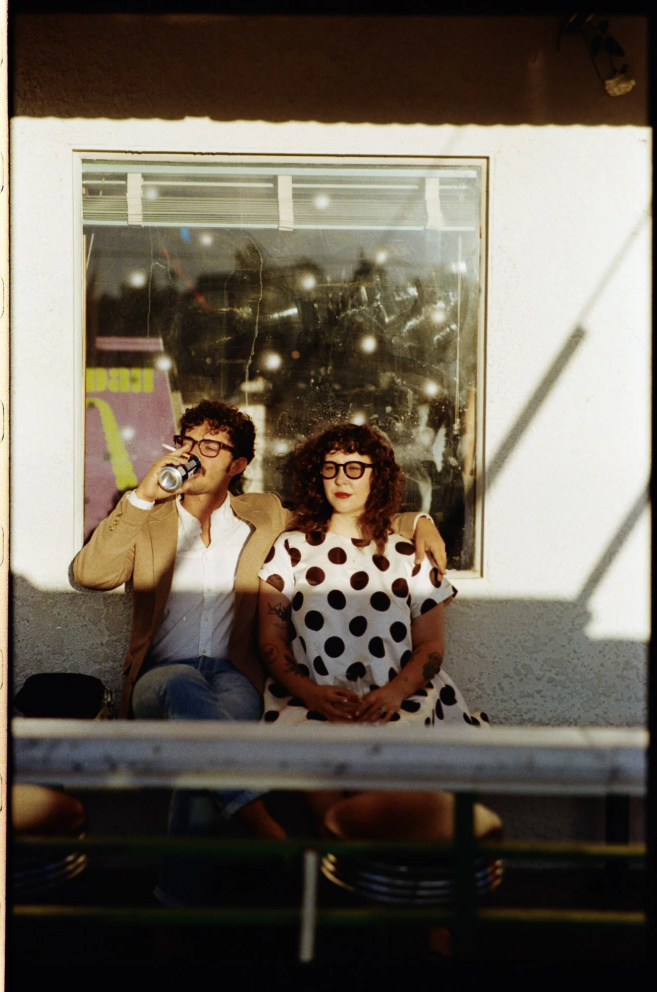 Couple sitting on a bench outside of a building, man has his arm wrapped around the girl. He is drinking a beer while holding a cigarette and she is wearing sunglasses looking off into the distance