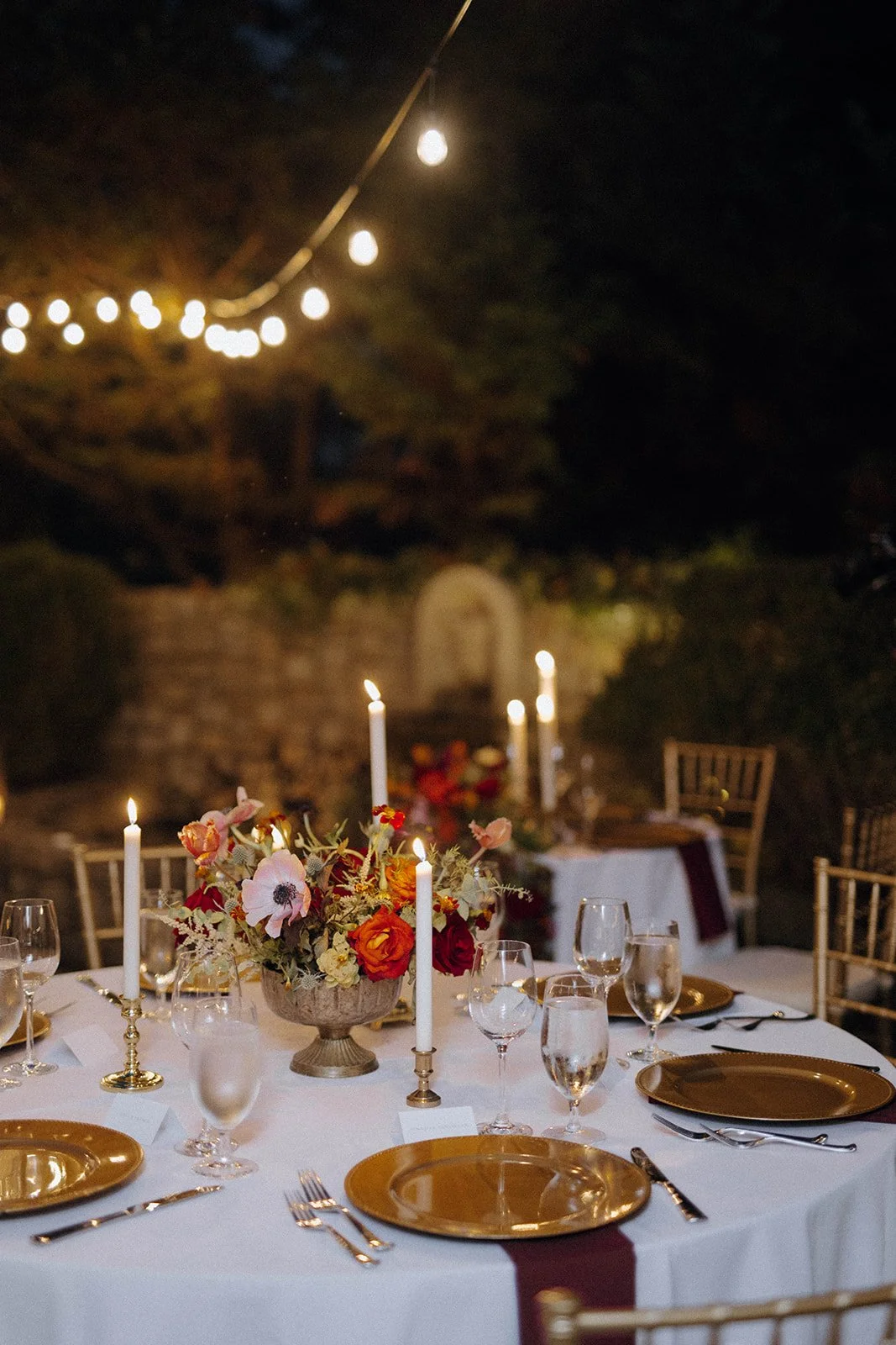 Night shot of an empty dining table featuring gold chargers, glasses, white candlesticks and poppies in silver urn arrangement