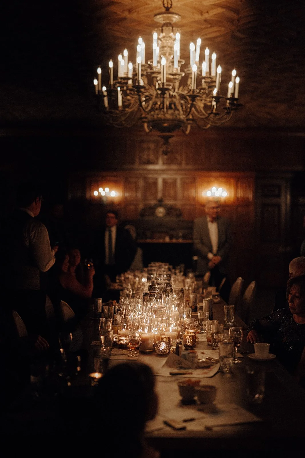 Dim lit dining room with large chandelier and dozens of glass pillar and votive candles down center of banquet-style table for wedding guests