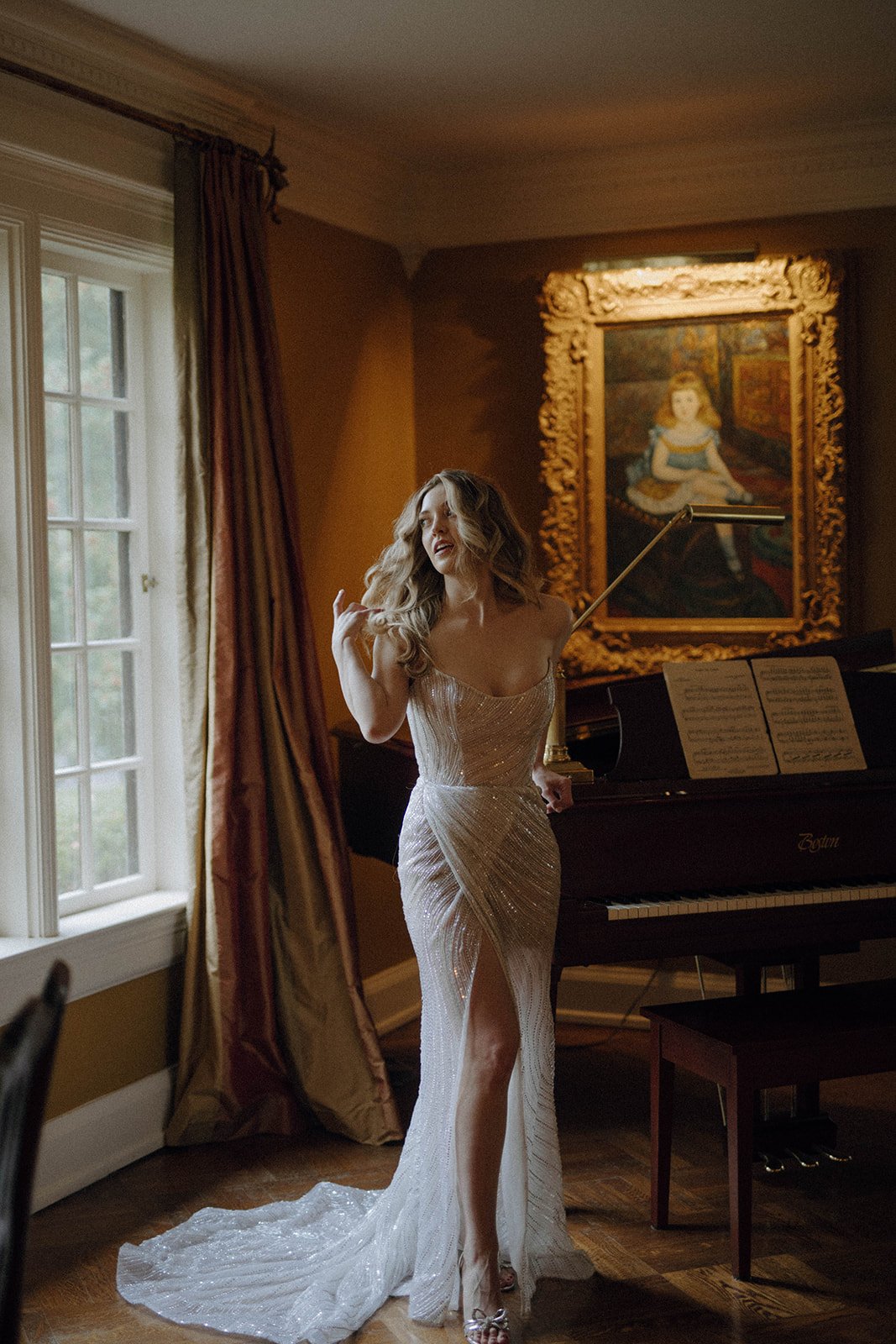 Bride wearing long lace gown flips her long wavy hair in a living room setting featuring piano, large picture window and gold framed portrait painting of little girl.