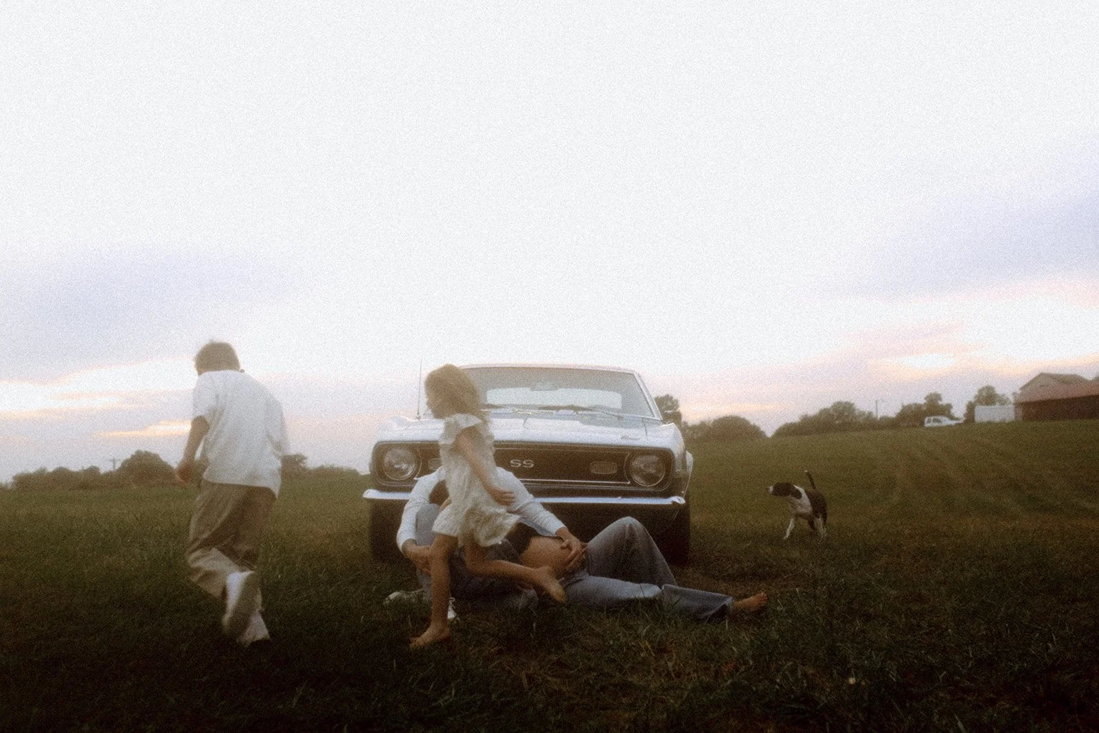 Family photo outside with children running around a vintage car with dog and parents watching them.