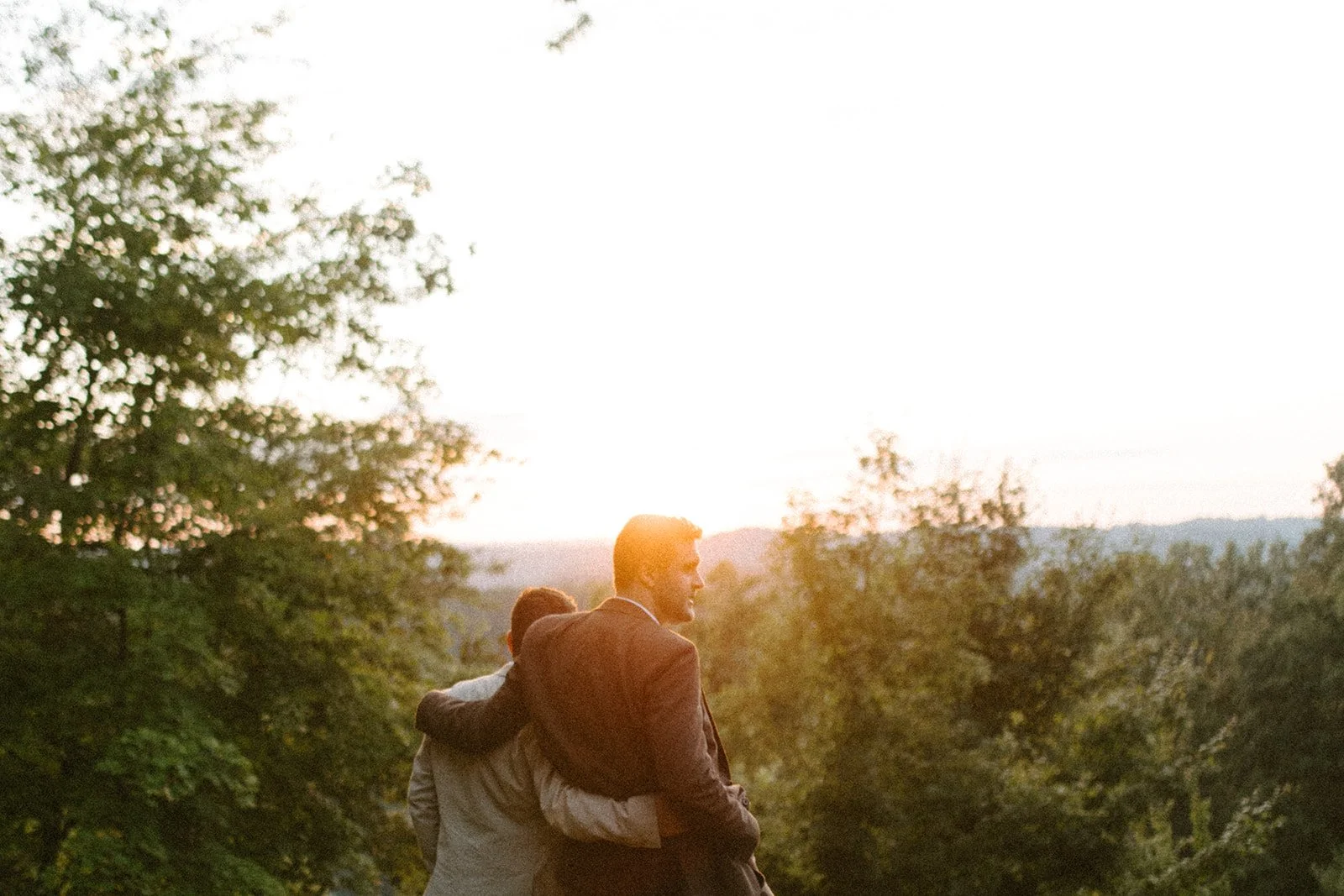 Shot from behind, a couple look over trees to sunset view with arms wrapped around each other