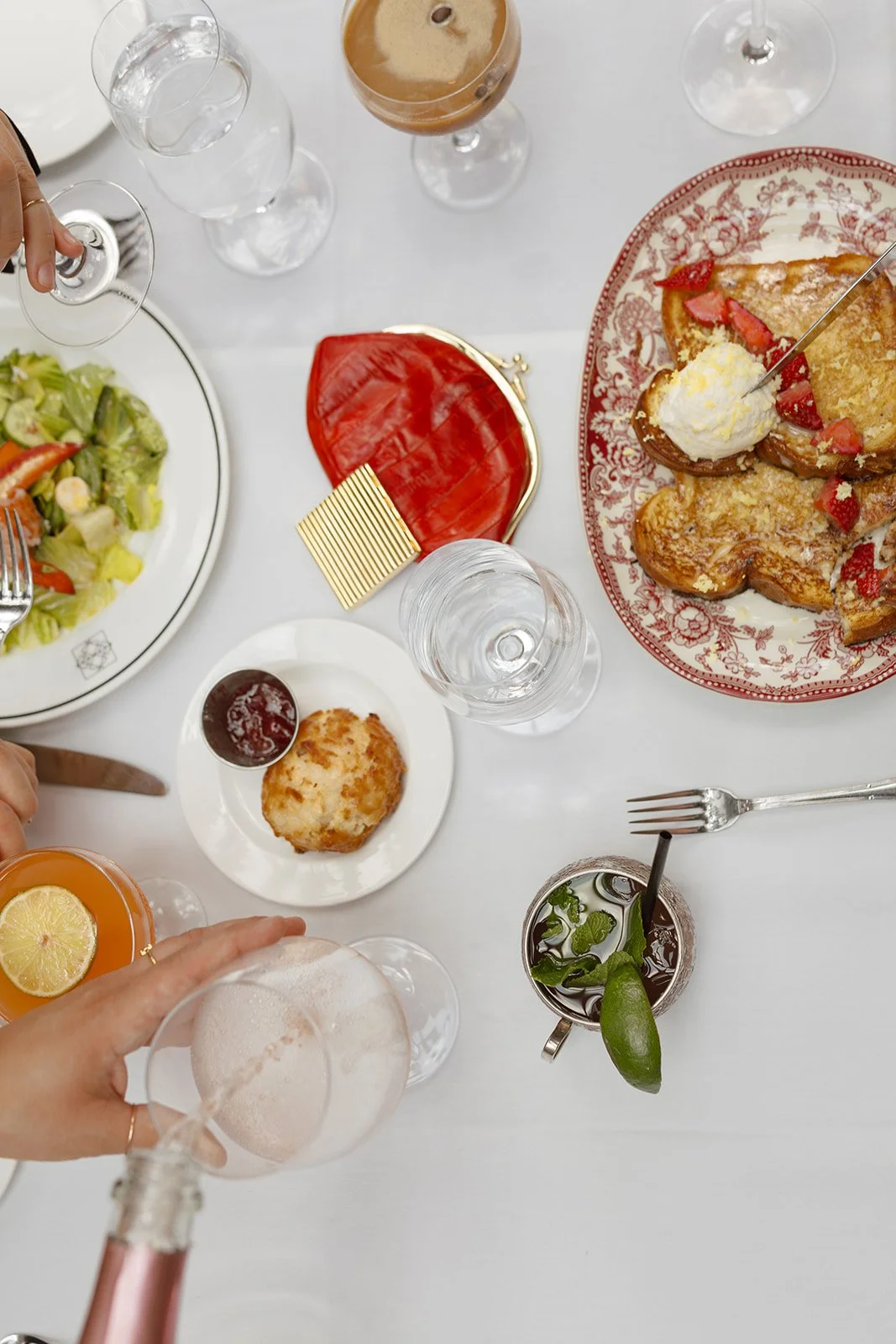 Overview of dining table featuring white tablecloth, water glasses and cocktails and some plates of food - leafy green salad, scone with jam, french toast with strawberries.