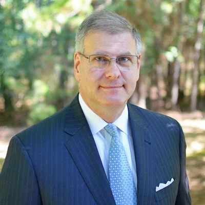Portrait of a middle-aged man in a navy pinstripe suit, light blue tie, wearing glasses, outdoors with trees.