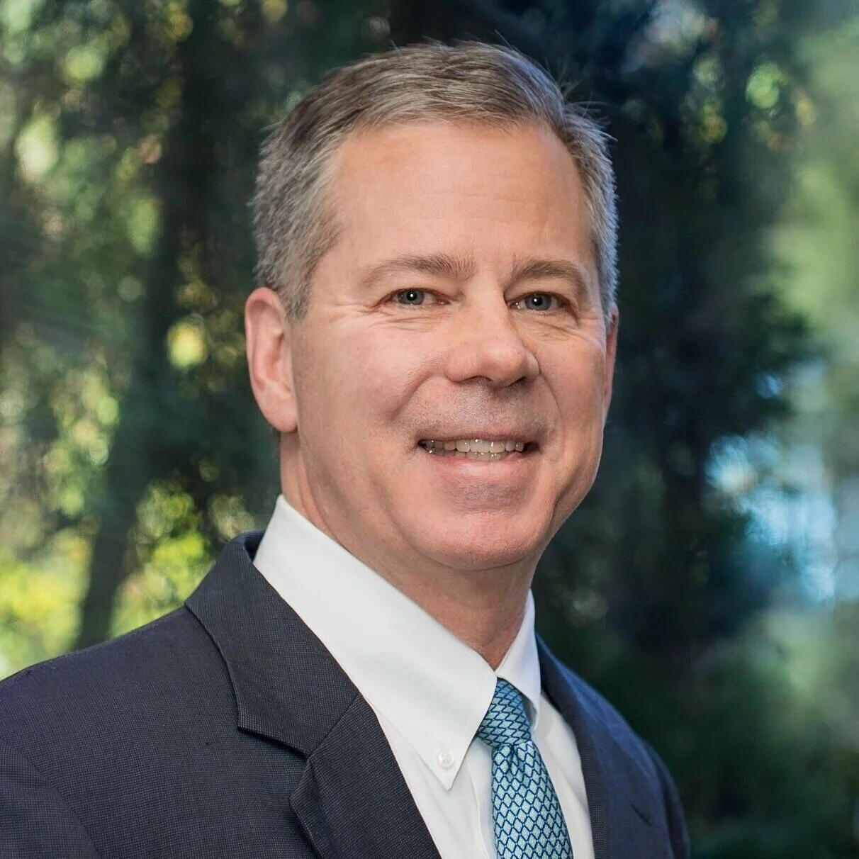 Portrait of a middle-aged man in a dark suit, white shirt, and patterned blue tie, smiling outdoors with blurred green trees in the background.