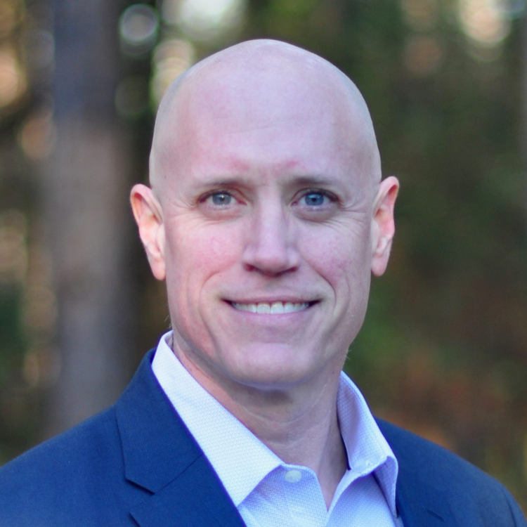 A man with a bald head and light skin wearing a blue suit jacket and a white collared shirt, smiling, outdoors with blurred trees in the background.