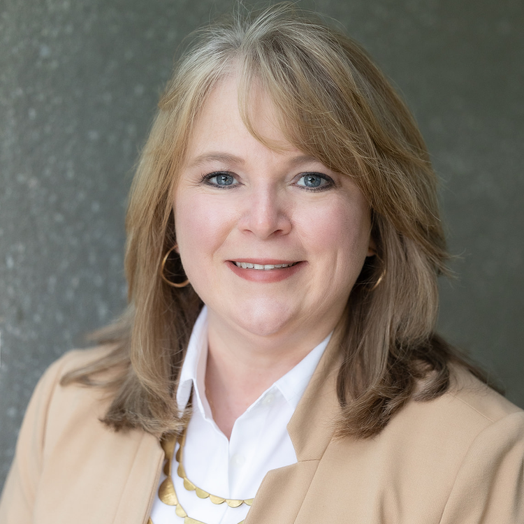 Close-up of a professional woman with shoulder-length light brown hair, blue eyes, wearing a beige blazer, white shirt, gold necklace, and small gold hoop earrings, smiling and looking at the camera against a gray textured background.