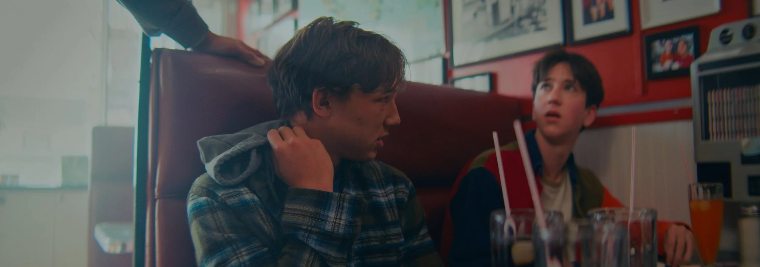 Two boys sitting at a restaurant booth, one adjusting his hoodie and the other looking at him with a surprised expression, with drinks on the table.