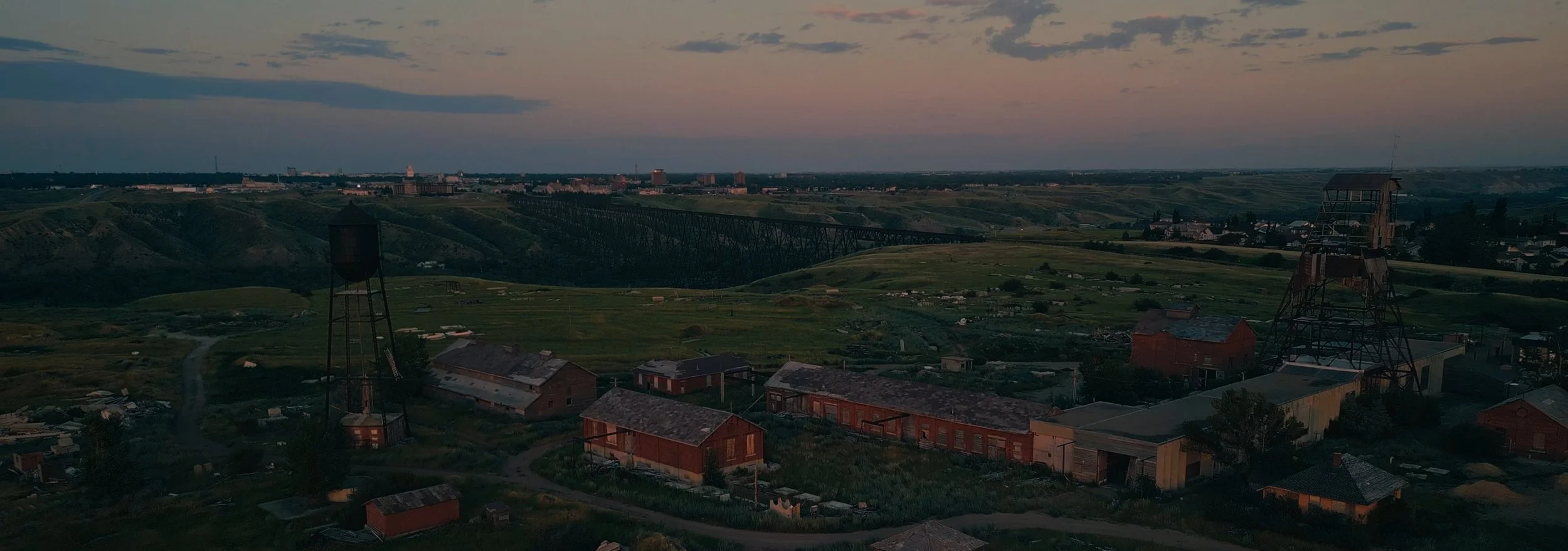 A landscape view of a rural area at dusk, featuring old wooden buildings, water towers, green hills, and a bridge in the distance under a cloudy sky.
