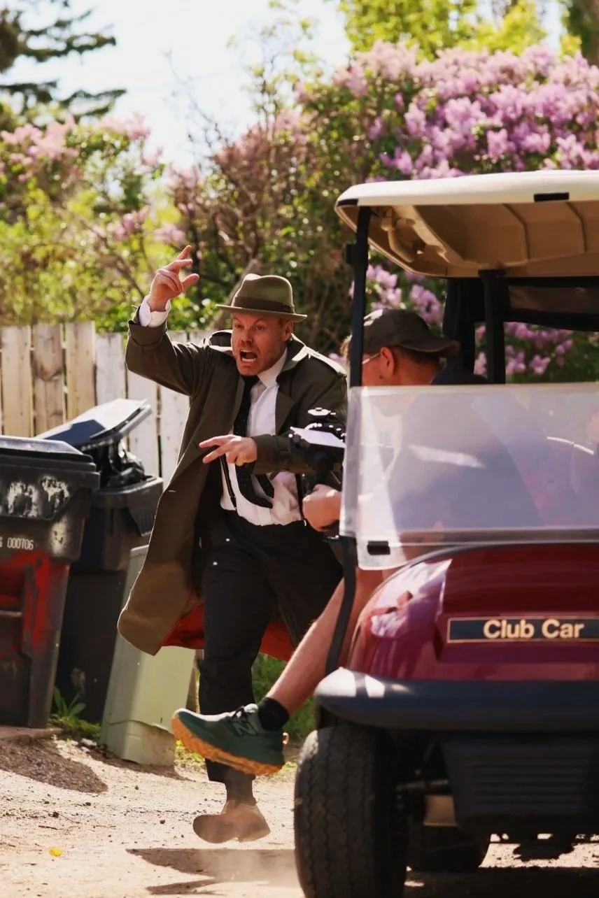 A man in a beige hat, beige coat, and white shirt appears angry or shouting, gesturing with his right hand, while standing near a golf cart labeled 'Club Car' during daytime. Another man, wearing a cap, is seated in the golf cart. The background features colorful pink and purple flowering trees and a wooden fence.