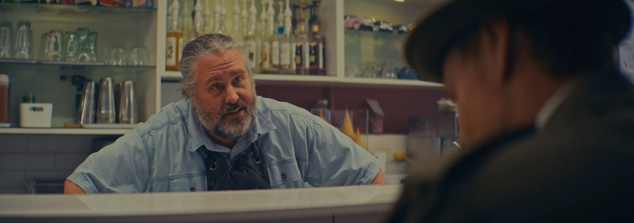 A man with gray hair and a beard, wearing a light blue shirt, working behind a counter in a store or cafe, engaged in conversation with a customer whose face is not visible.