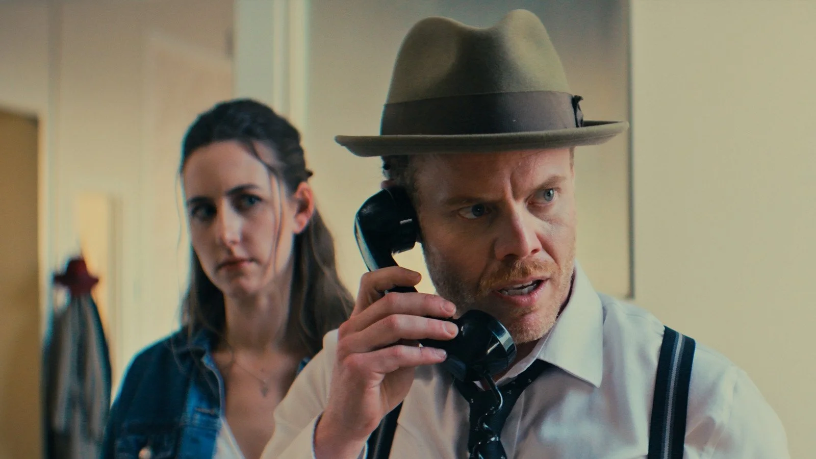 A man wearing a fedora talking on a vintage telephone with a woman standing behind him in an office setting.