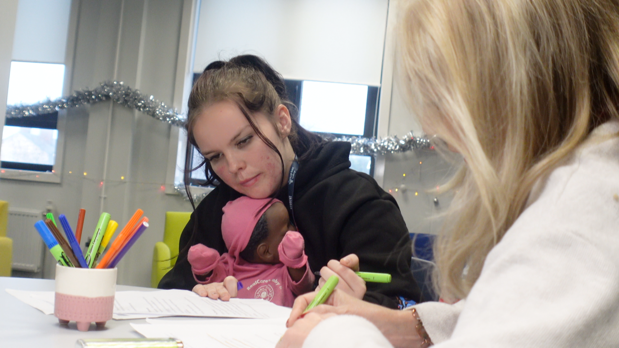 A woman with dark hair and a black hoodie holds a small child dressed in pink with a pink hat. They are seated at a table with papers and colorful markers, and another woman with blonde hair is also at the table. The scene appears to be in a decorated indoor setting with windows and holiday decorations.