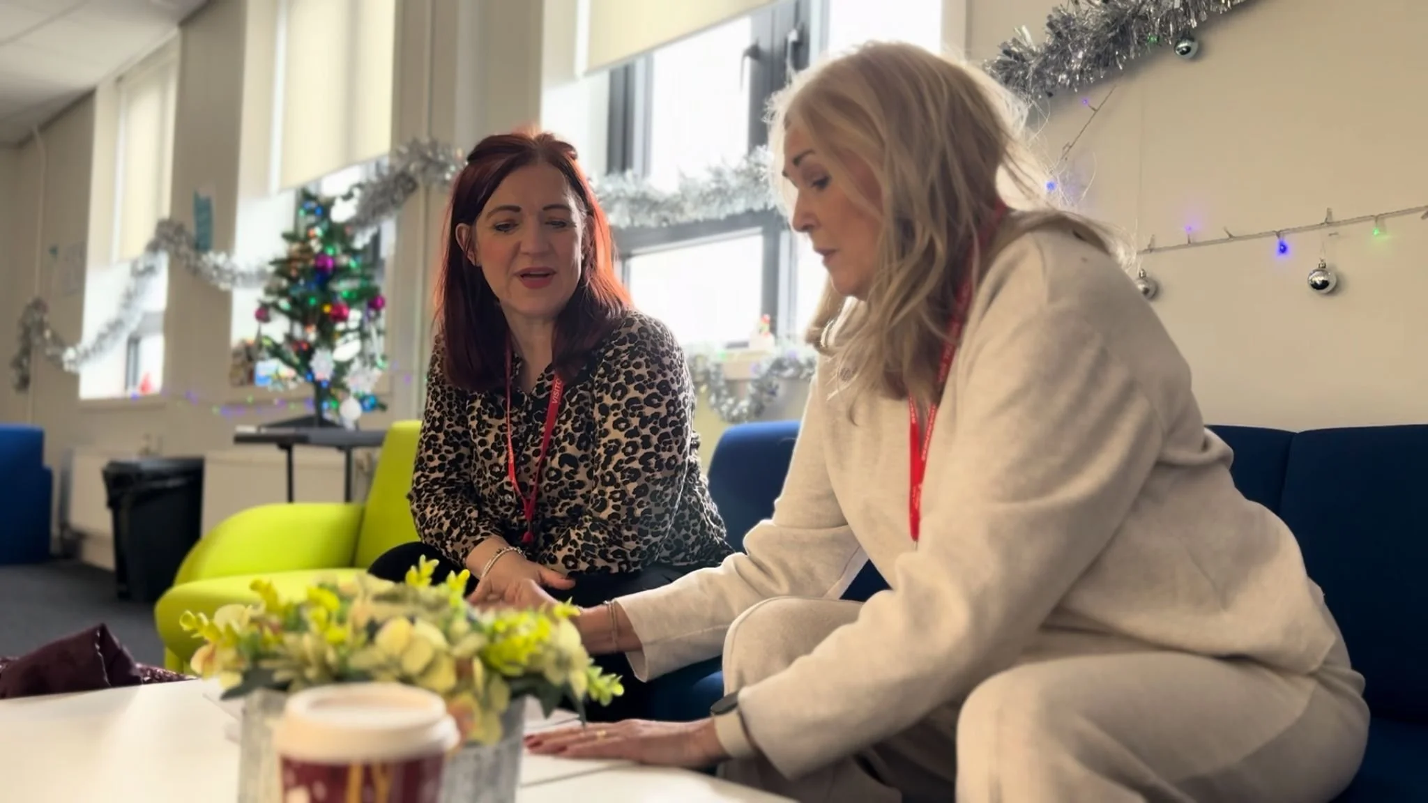 Two women sitting on a couch in a decorated room, with a Christmas tree and holiday decorations in the background, having a conversation.