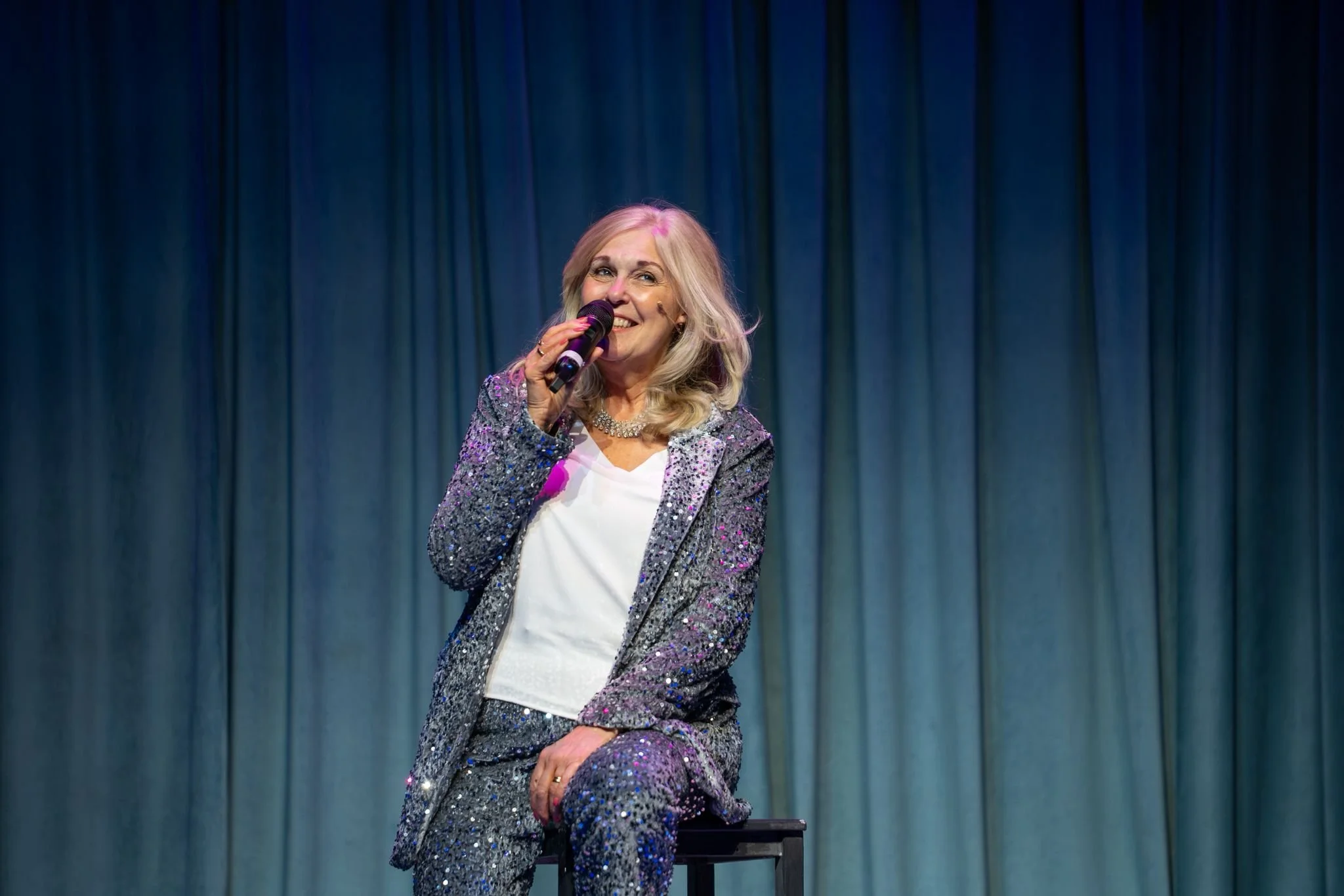 A woman wearing a sparkly suit, holding a microphone, sitting on a stool on stage with a blue curtain background, smiling.