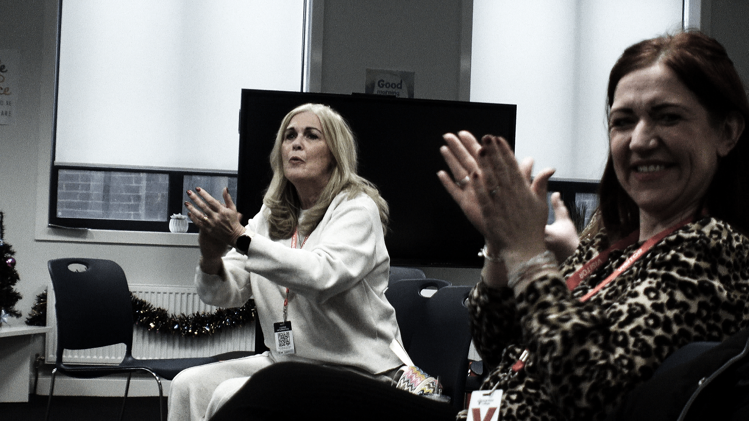 Two women sitting in a room, clapping and listening. The woman on the left has long blonde hair and is wearing a white blazer, the woman on the right has short red hair and is wearing a leopard print top. There is a large monitor and Christmas decorations in the background.