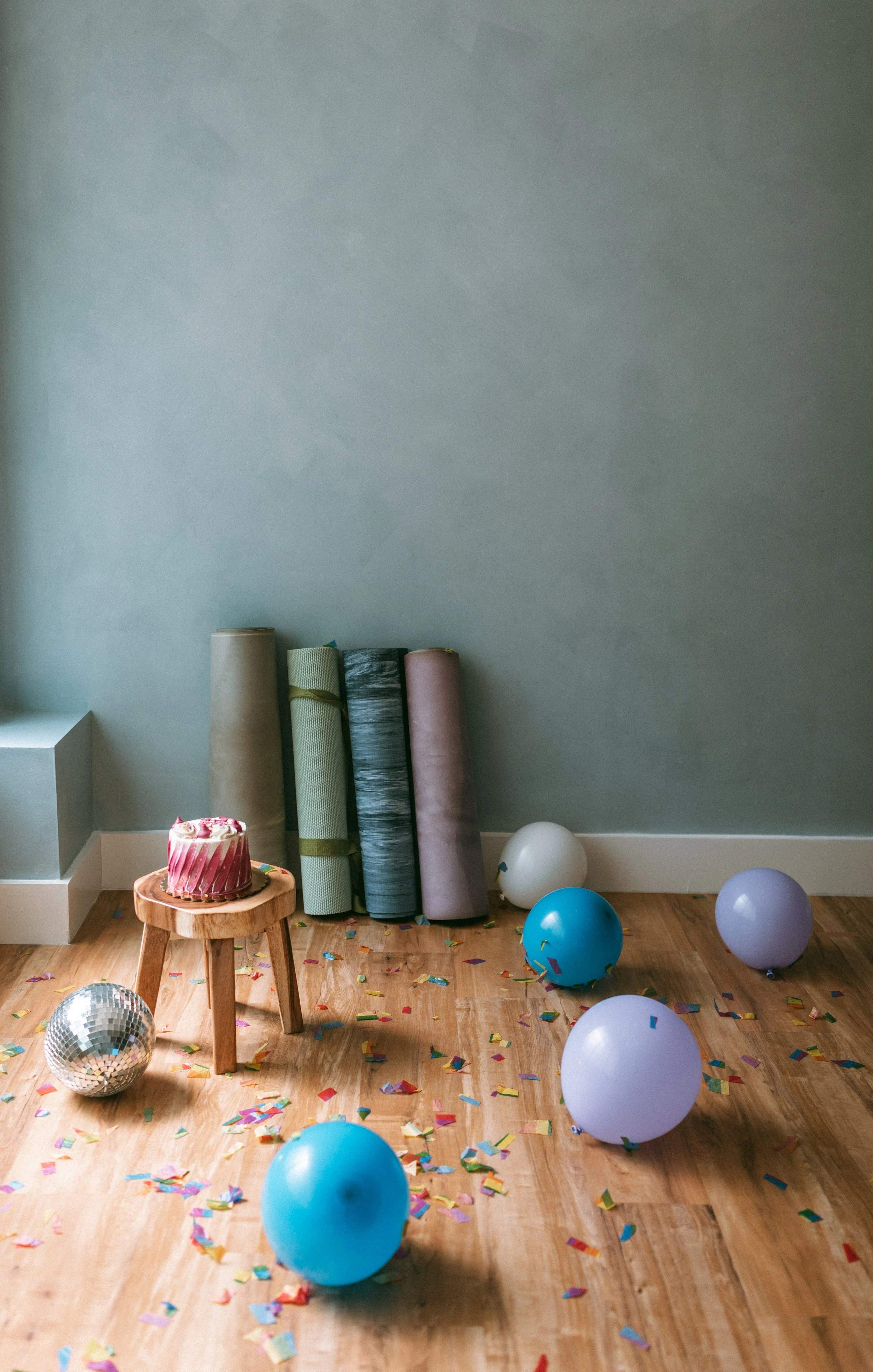 Room decorated for celebration with confetti, balloons, a small cake on a wooden stool, and yoga mats leaning against the wall.