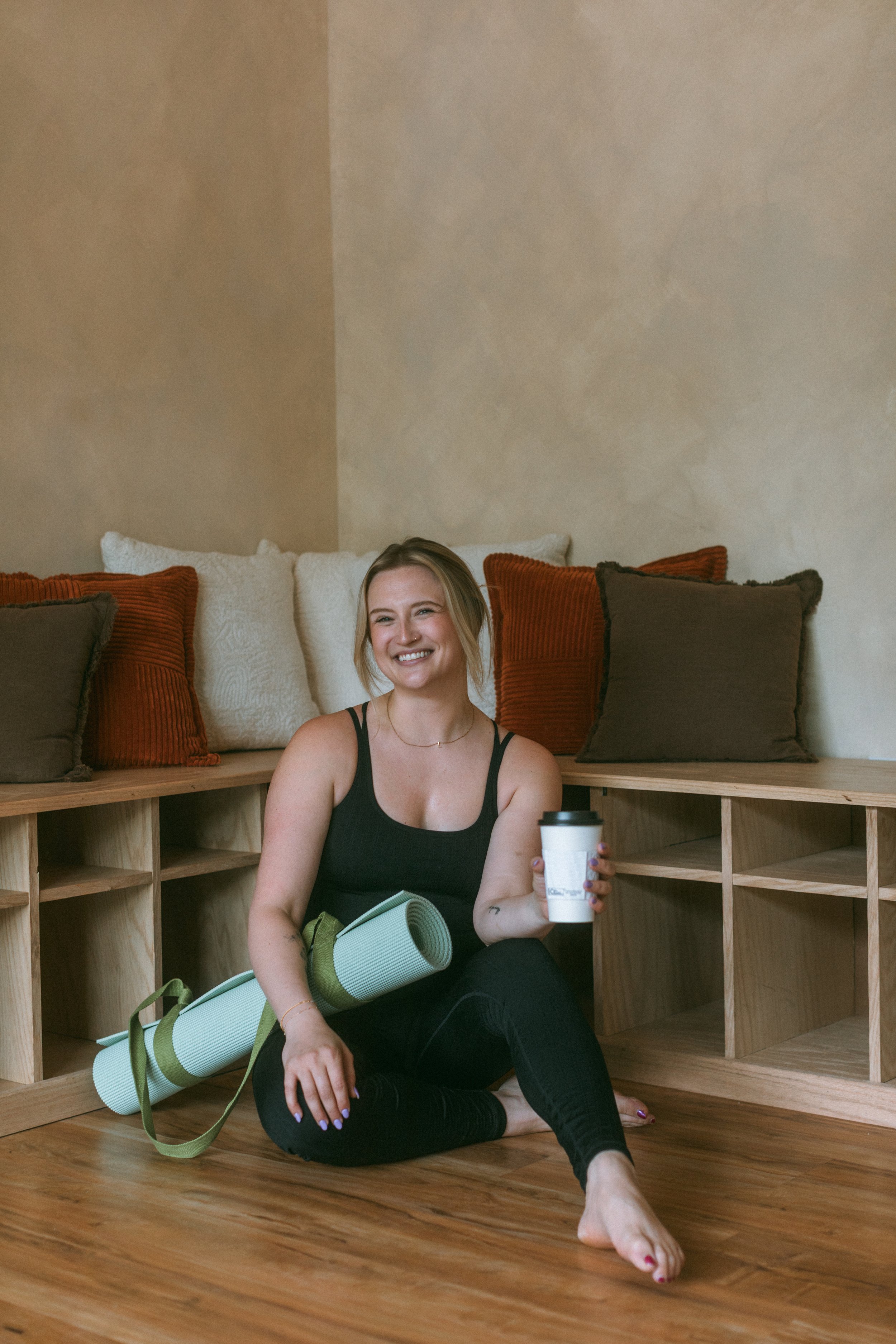 A young woman sitting on the wooden floor, holding a coffee cup, with a yoga mat on her lap, smiling and looking happy in a cozy room.