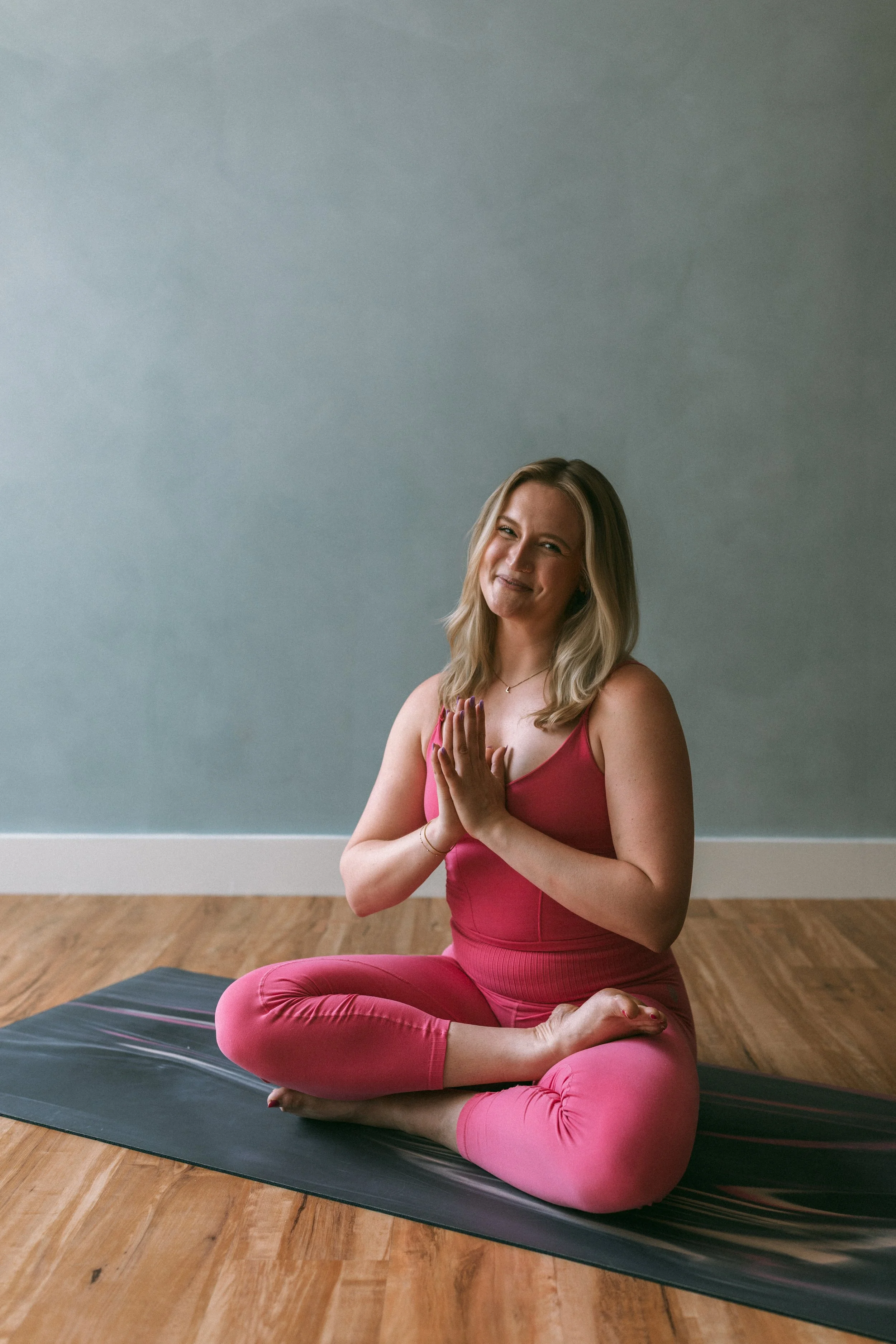 A woman in a pink yoga outfit sitting cross-legged on a black yoga mat indoors, smiling and doing a prayer pose with her hands together.
