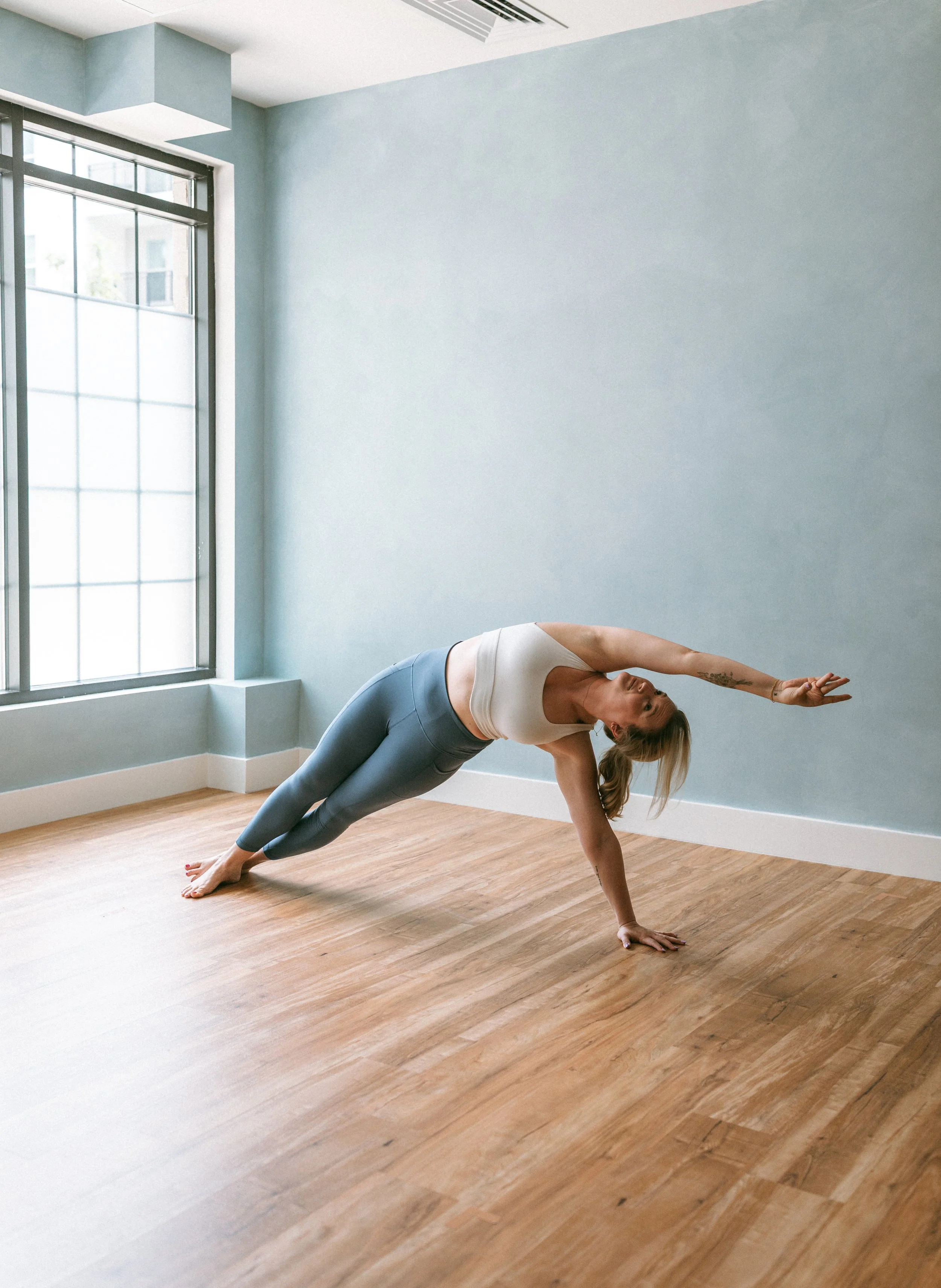 A woman practicing yoga indoors, balancing on her right hand and feet, with her left arm extended upward. She is on a wooden floor next to a large window with grid panes, in a room with light blue walls.