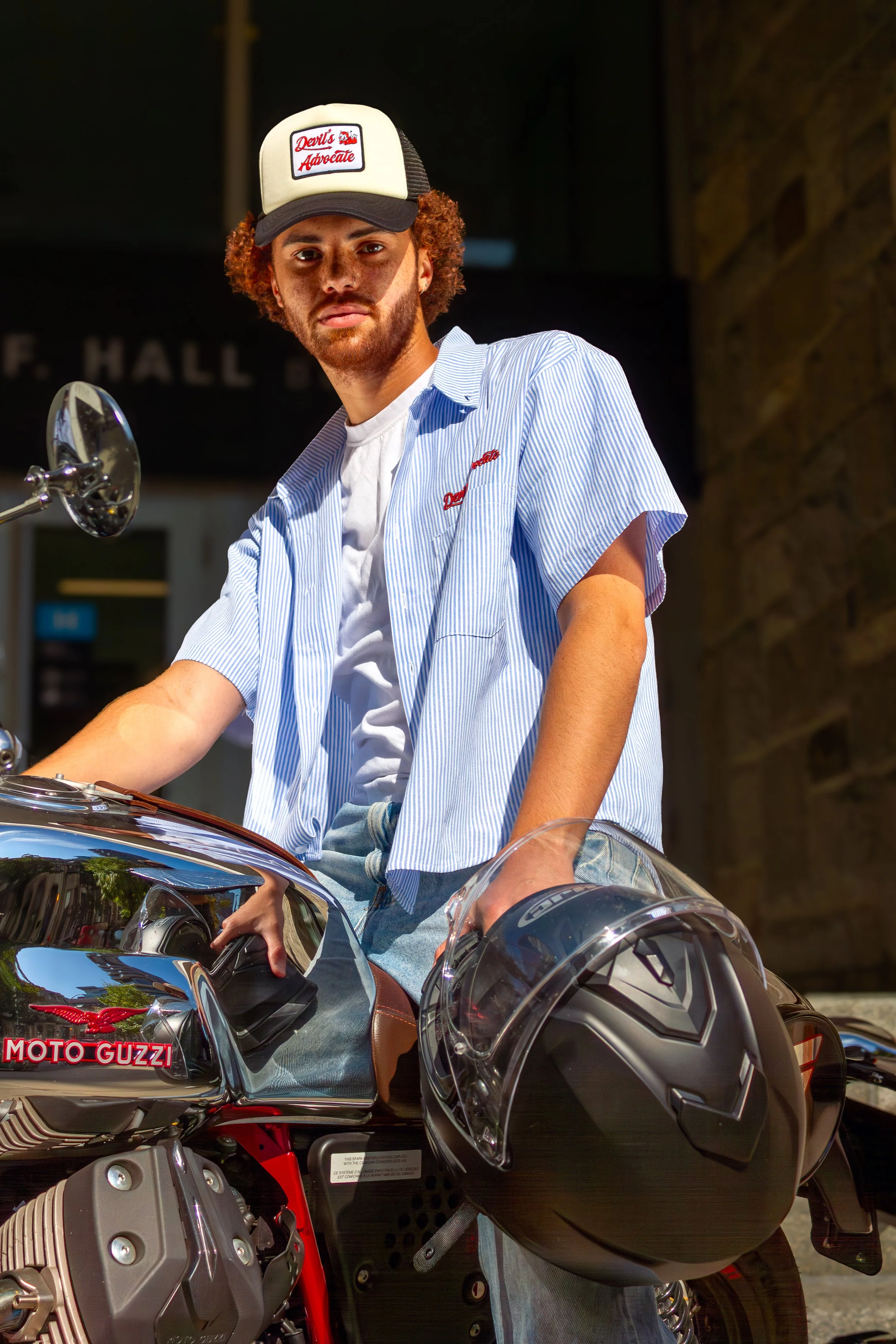 A young man with curly hair and a beard sitting on a motorcycle, wearing a trucker hat, a blue and white button up striped shirt, and casual jeans, holding a black helmet with a visor.
