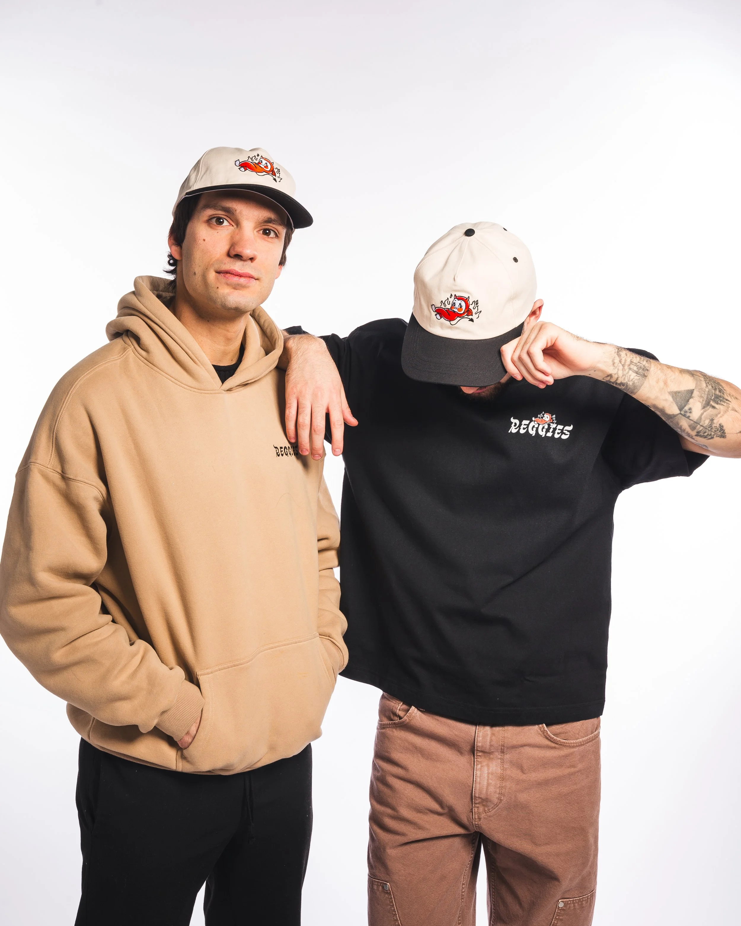 Two men wearing matching beige and black baseball caps, standing against a white background. The man on the left wears a beige hoodie with the same logo, while the man on the right wears a black T-shirt with the logo and has tattoos on his arm. The m