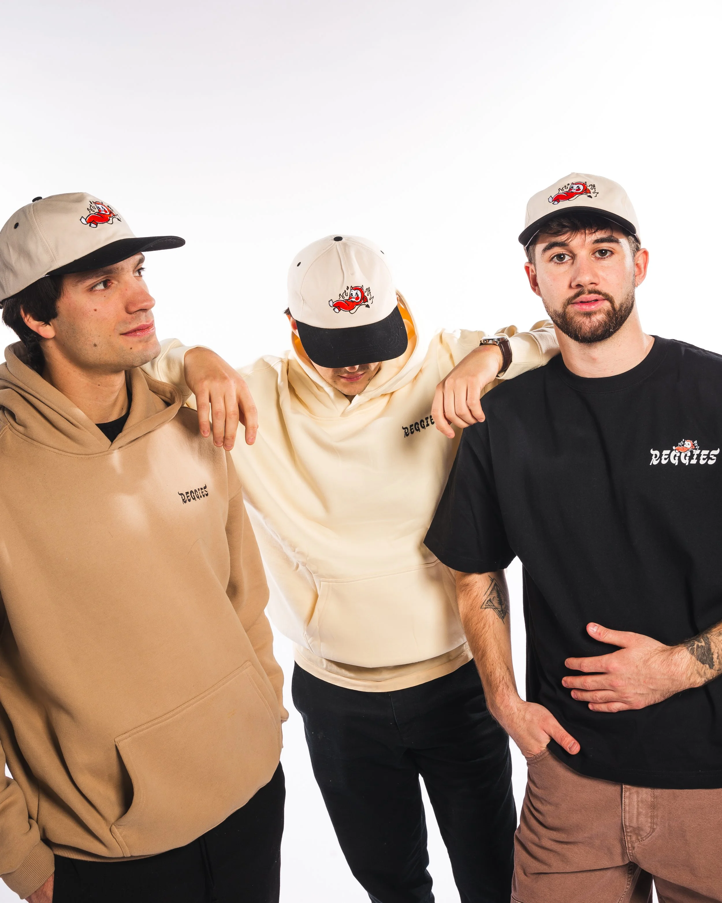 Three young men in casual clothing and beige baseball caps with a red devil logo, standing shoulder to shoulder with arms around each other, posing against a plain white background.