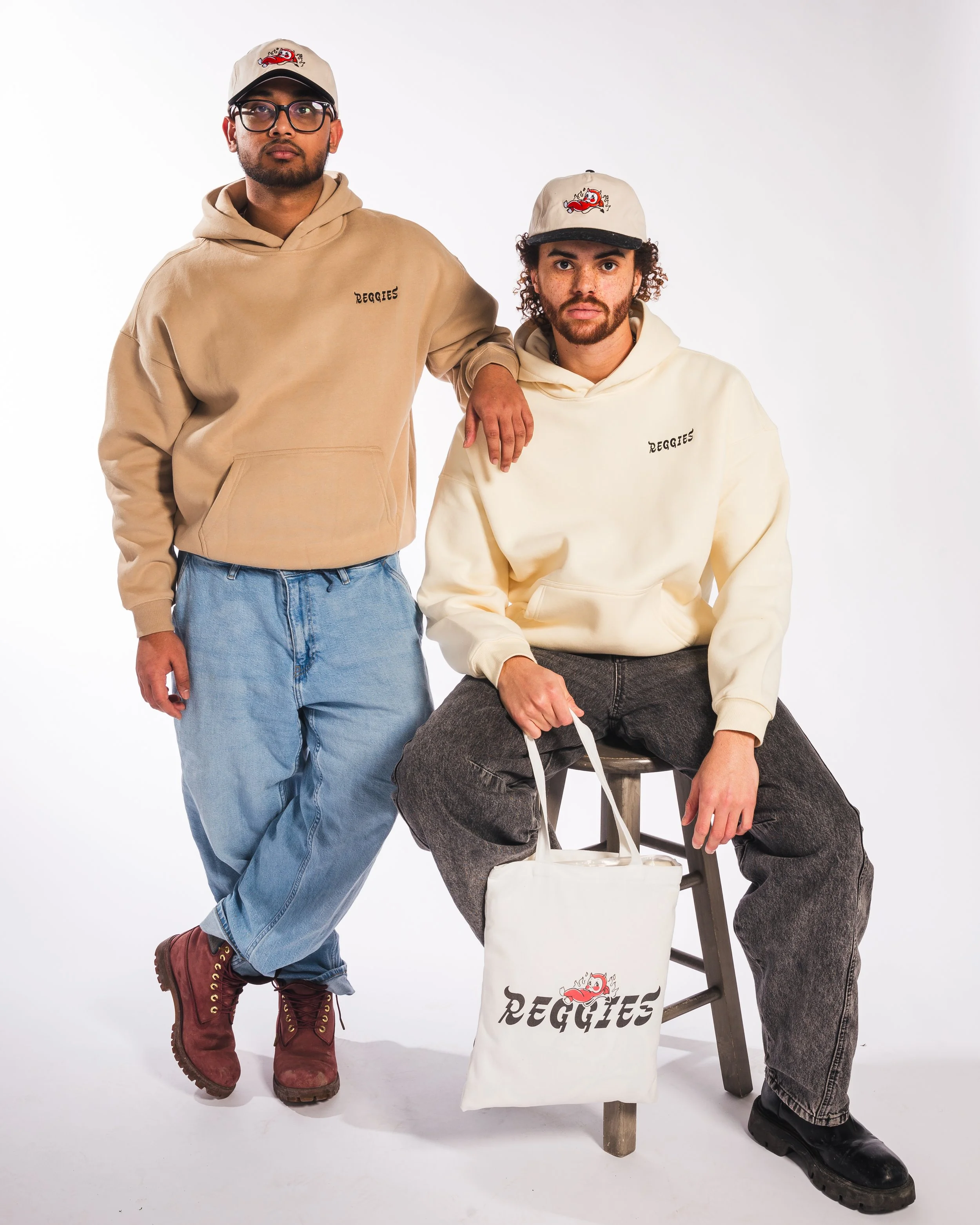 Two men wearing beige Reggie's hoodies and caps, are posing with a white Reggie's tote bag. One man is standing with his hand on the other's shoulder. The other is sitting on a stool. The background is plain white.