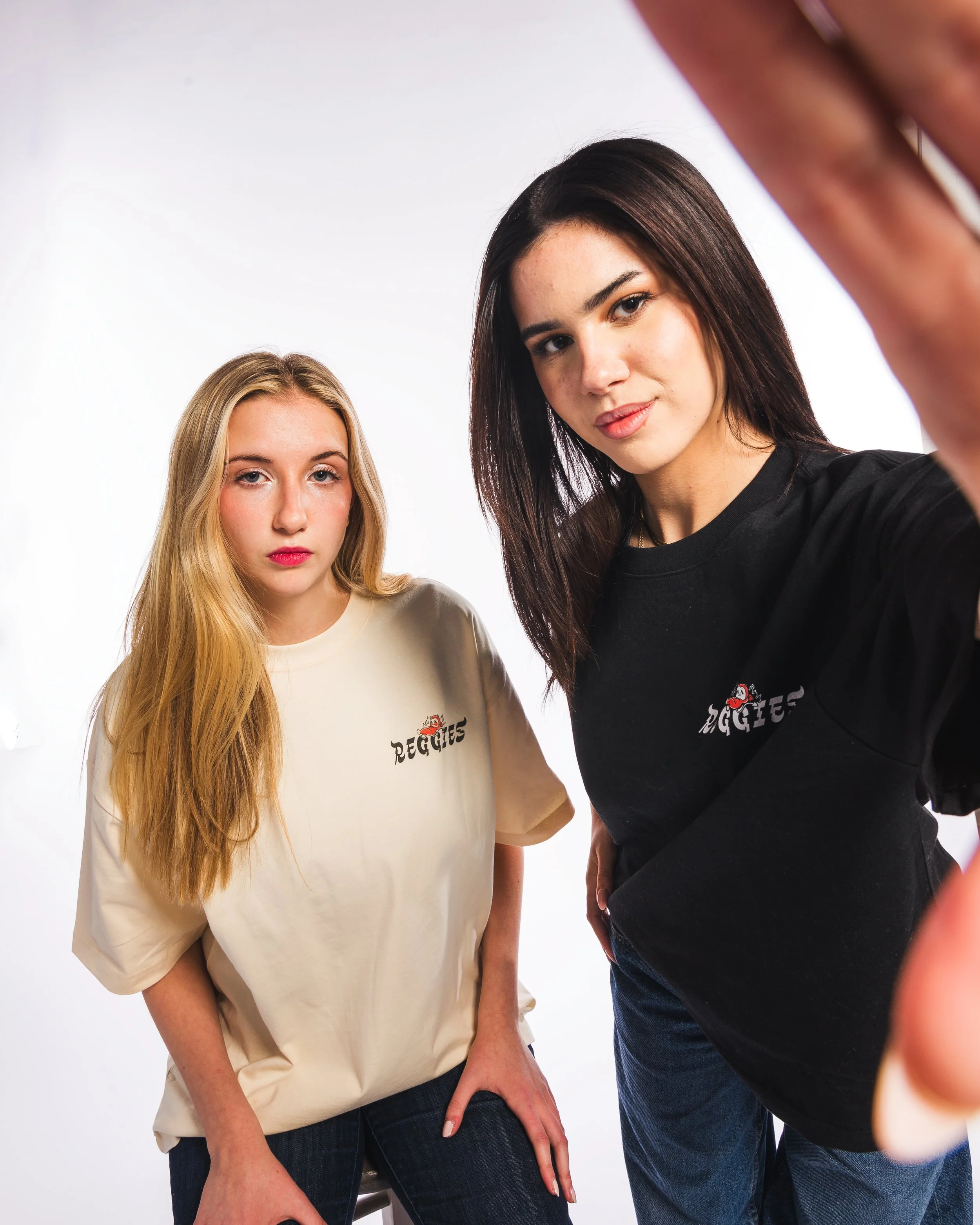 Two women taking a selfie against a white background, one with long blonde hair wearing a beige sweatshirt, and the other with long dark hair wearing a black t-shirt, both with the Reggie's logo.