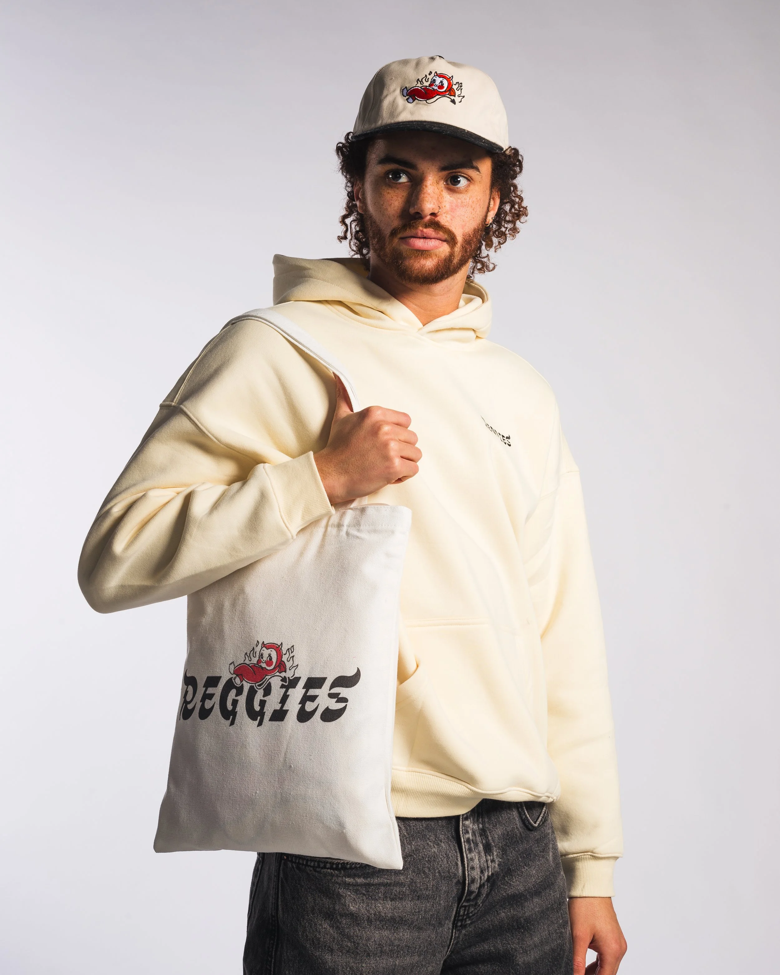 A young man with curly hair and a beard wearing a white hoodie, a beige cap with a cartoon devil logo, and carrying a tote bag with the same devil logo and the text 'REGGIES'.