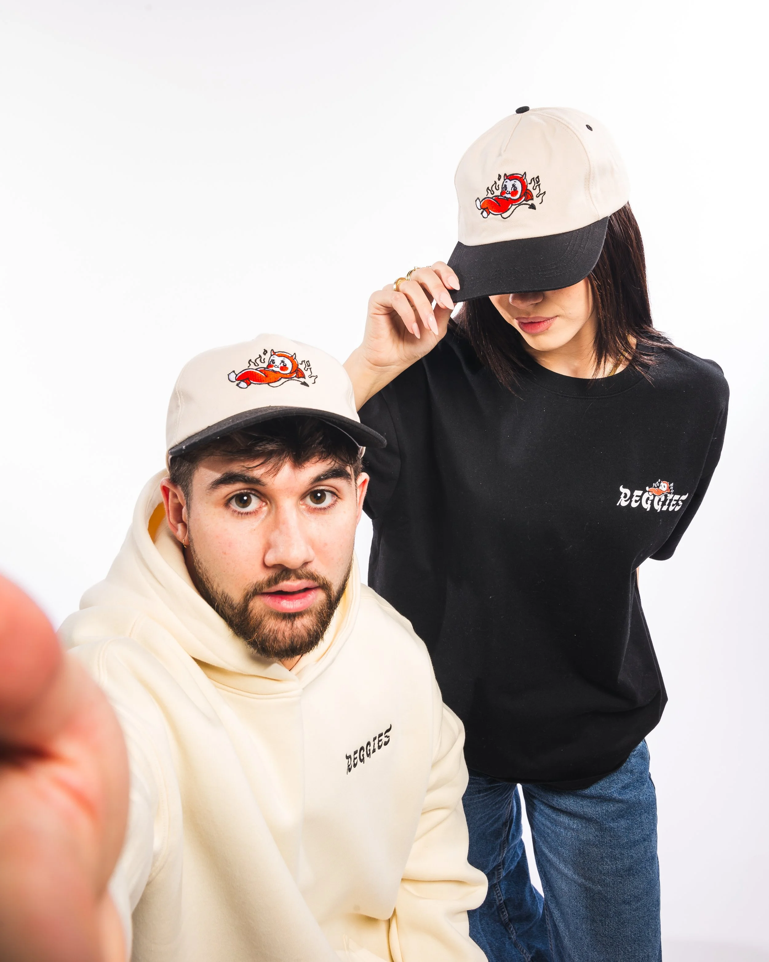 Man and woman wearing matching beige and black caps with a cartoon devil logo, both dressed in black and beige hoodies with 'REGGIES' text, posing against a white background.