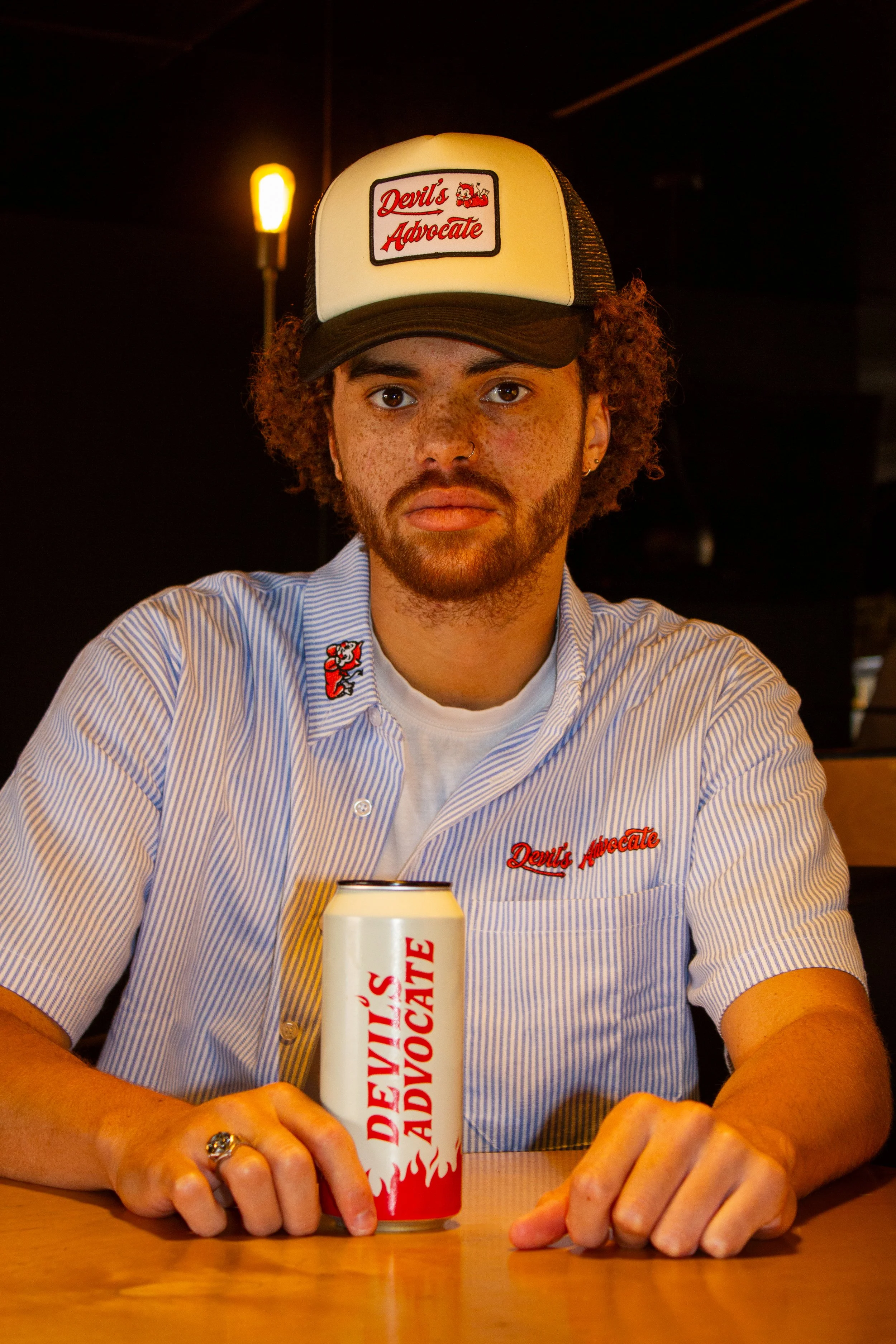 A young man with curly hair and freckles sits at a table inside a dimly lit restaurant. He wears a white and blue striped short-sleeve shirt with an embroidered patch and a white trucker hat with a patch that reads 'Devil's Advocate'. On the table in