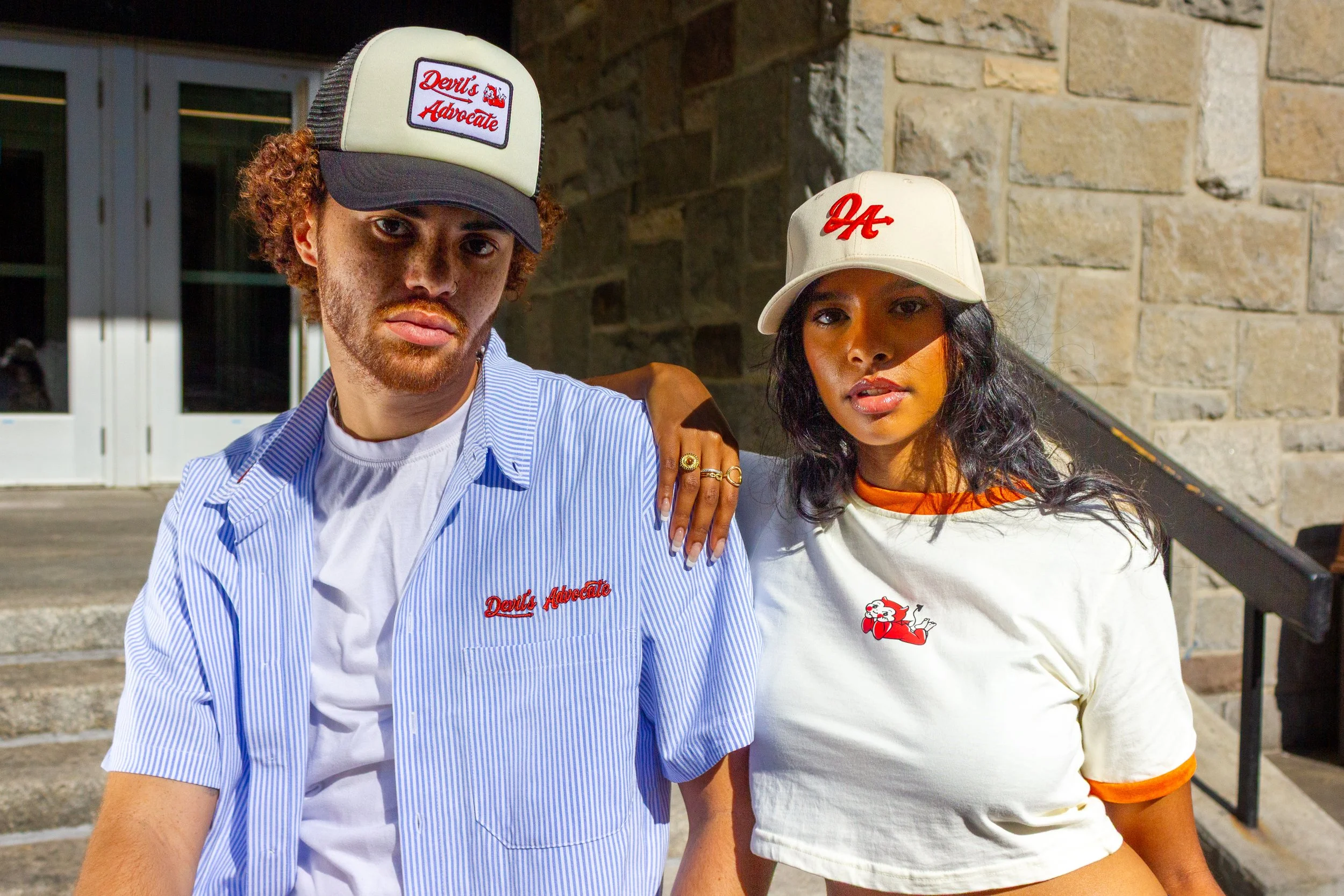 Two young adults, a man and a woman, standing close together on outdoor stairs, wearing casual outfits with caps that have red text and logos. The man has curly red hair and a beard, wearing a striped button-up shirt over a white t-shirt. The woman h
